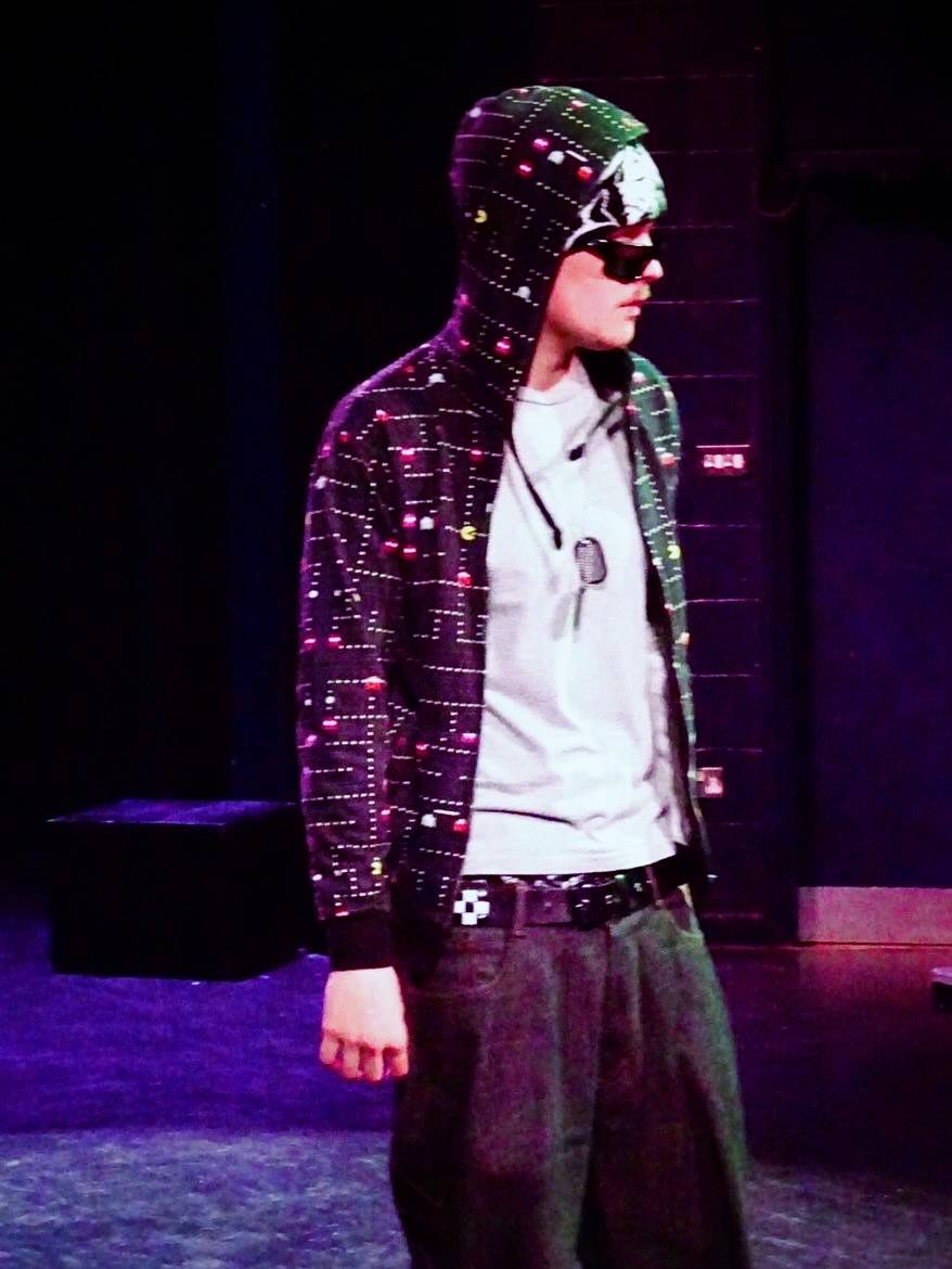 Performer on a dark stage wearing a hooded, LED-pattern jacket and sunglasses, facing to the right.