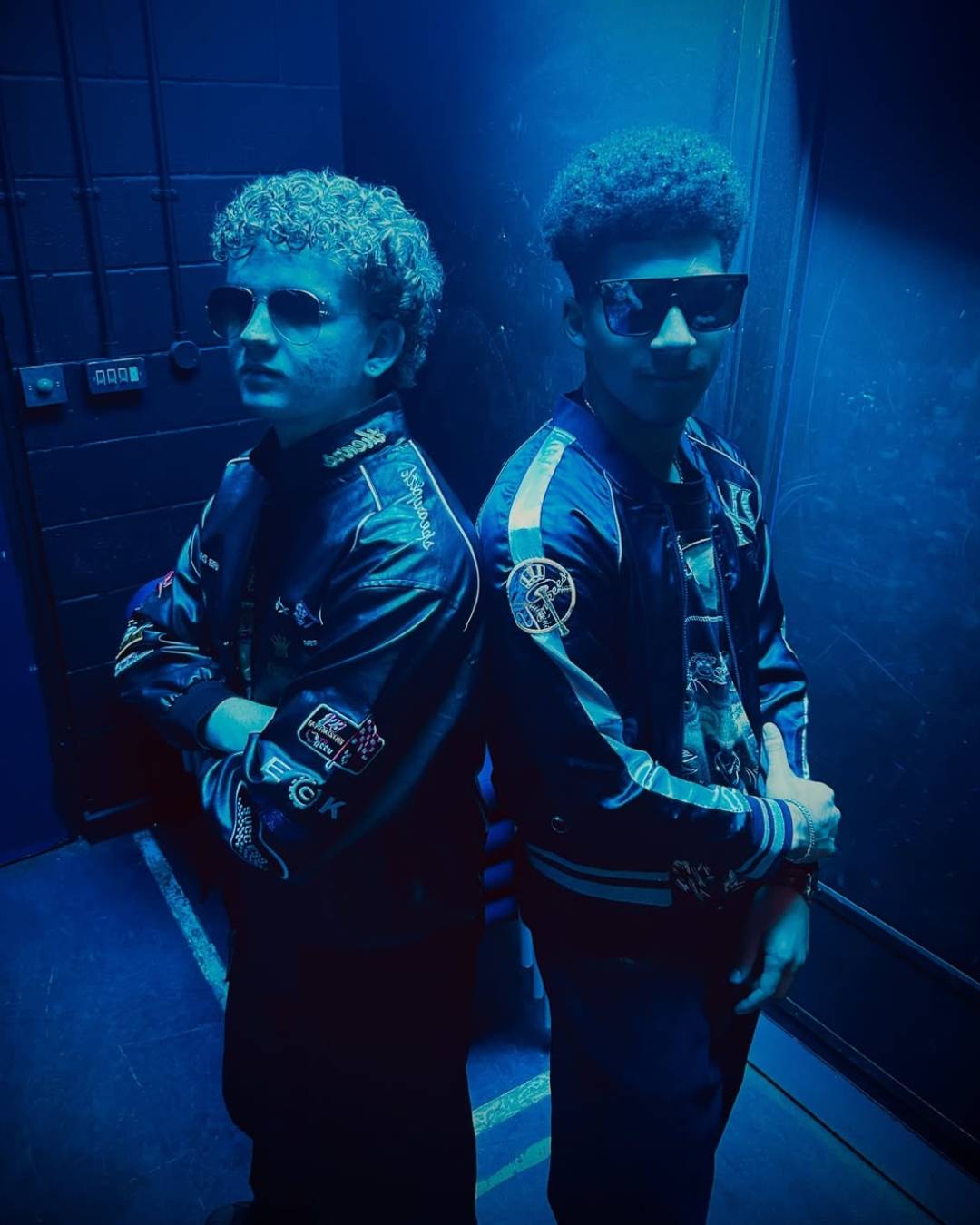 Two young men pose back-to-back in blue lighting, wearing bomber jackets with patches and sunglasses in a dim hallway.