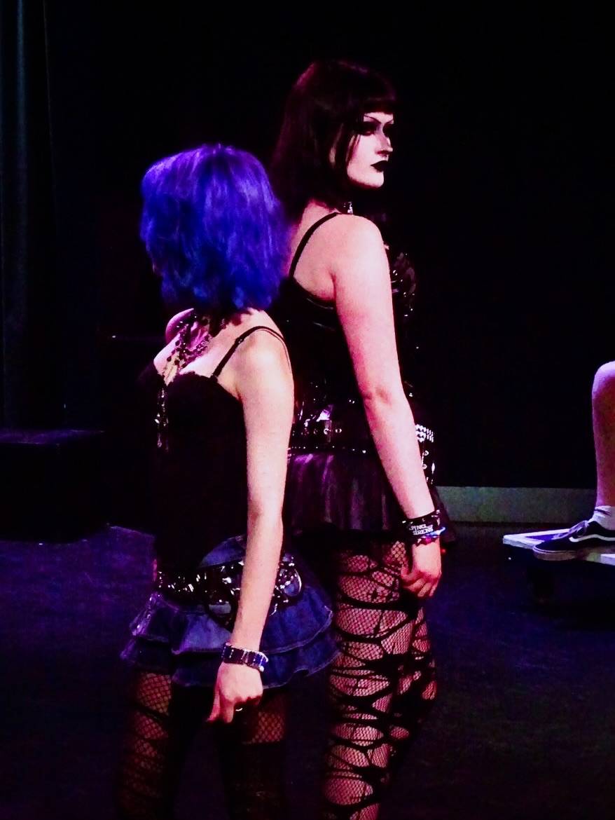 Two performers stand back-to-back on a dark stage, one with bright blue hair and a ruffled denim skirt with fishnet tights.
