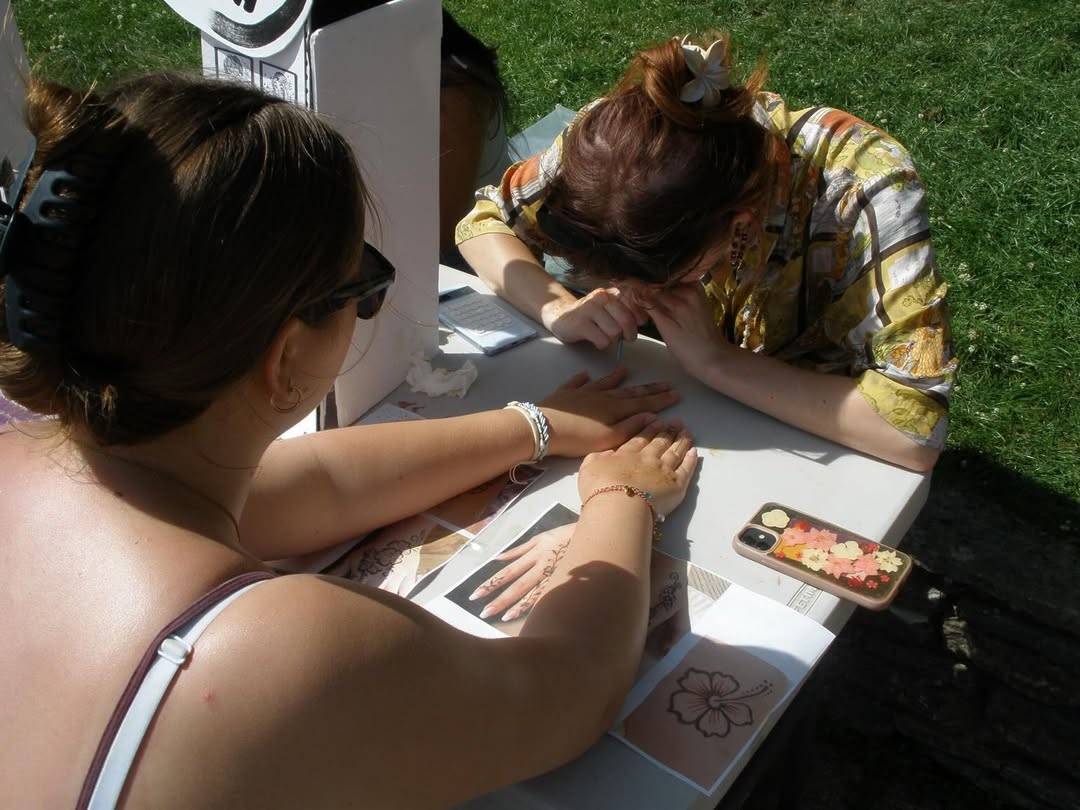 Two women share a manicure outdoors on a sunny day; one nails the other’s hand at a white table with flowers-patterned papers nearby and a floral phone case nearby.