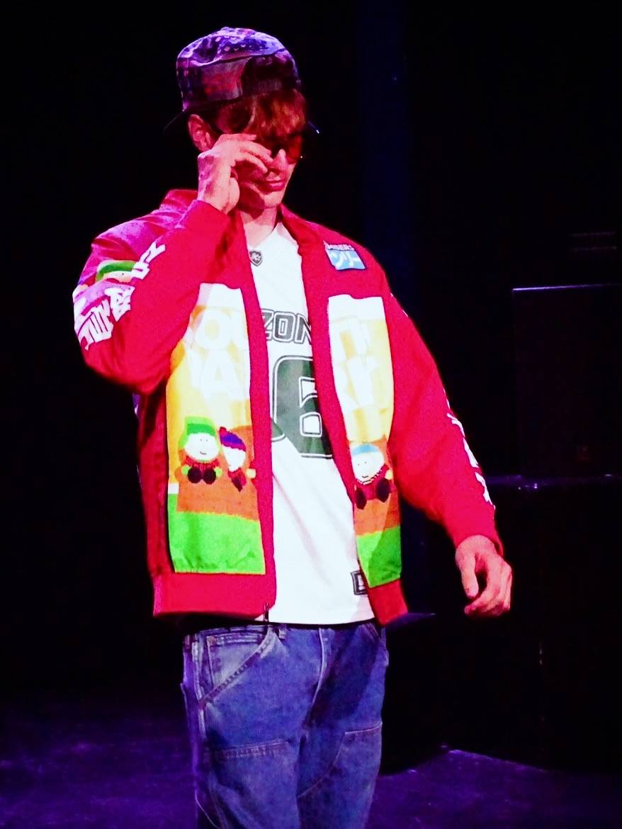 Male performer on a stage wearing a red festive jacket with cartoon patches, white shirt, blue jeans, and a cap, adjusting his sunglasses.