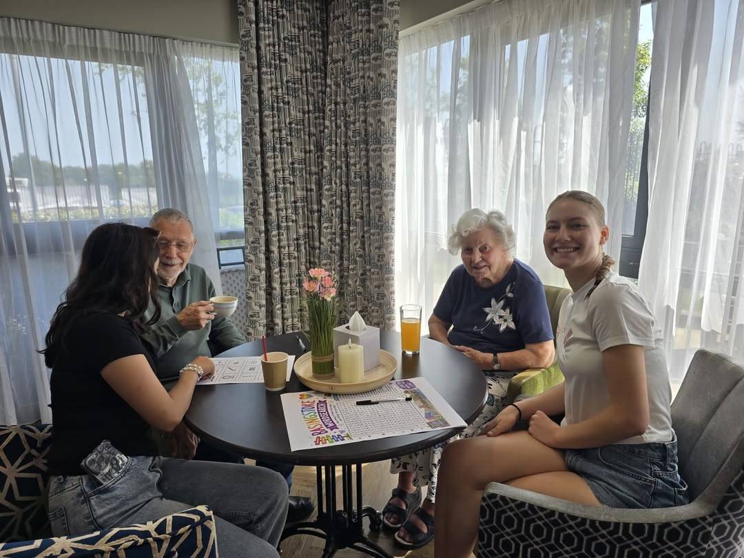 Four people sit around a round table in a bright lounge, sharing coffee and conversation near sheer curtains and a vase of pink flowers on the table.