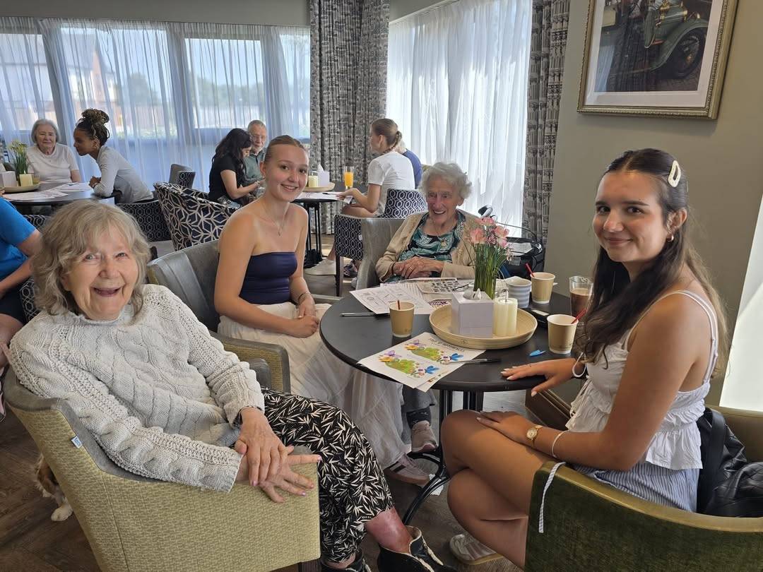Four women of varying ages sit around a round cafe table, smiling, with drinks and a vase of flowers on the table in a bright room.