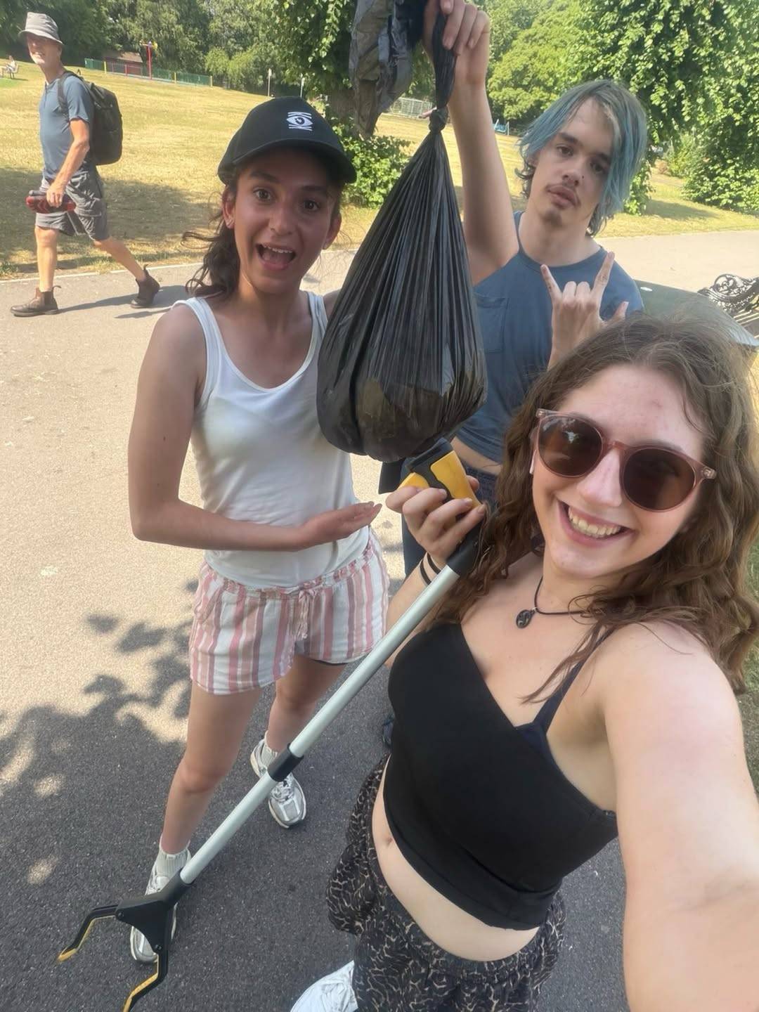 Three friends in a park pose for a photo, holding a large black trash bag with a litter picker, smiling at the camera.