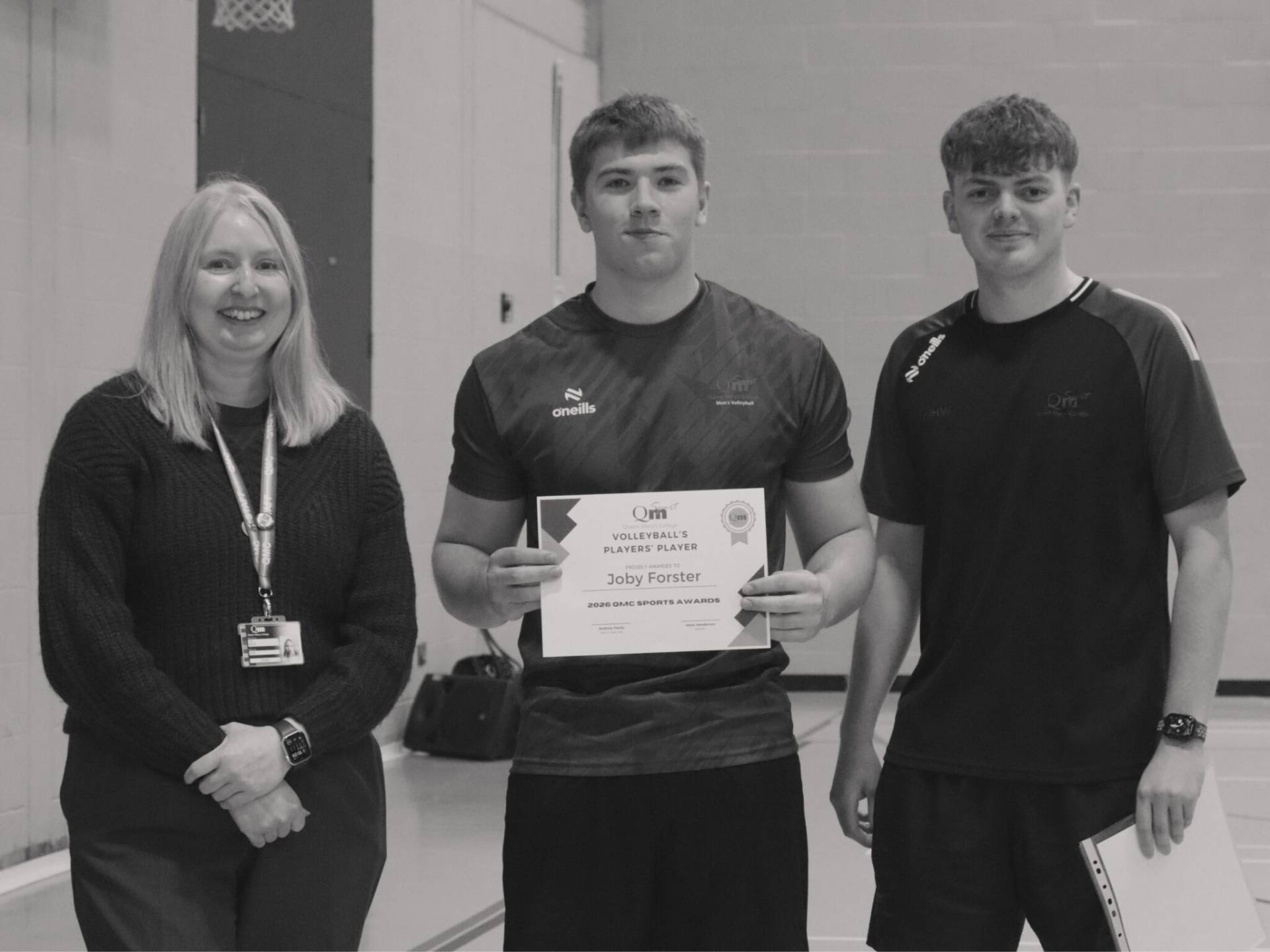Three people stand in a gym: a smiling woman on the left, and two men in the center and right; the middle man holds a certificate.