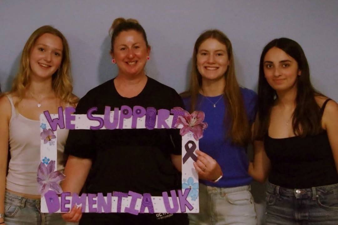 Four women smiling, holding a purple frame that says 'WE SUPPORT DEMENTIA UK'.