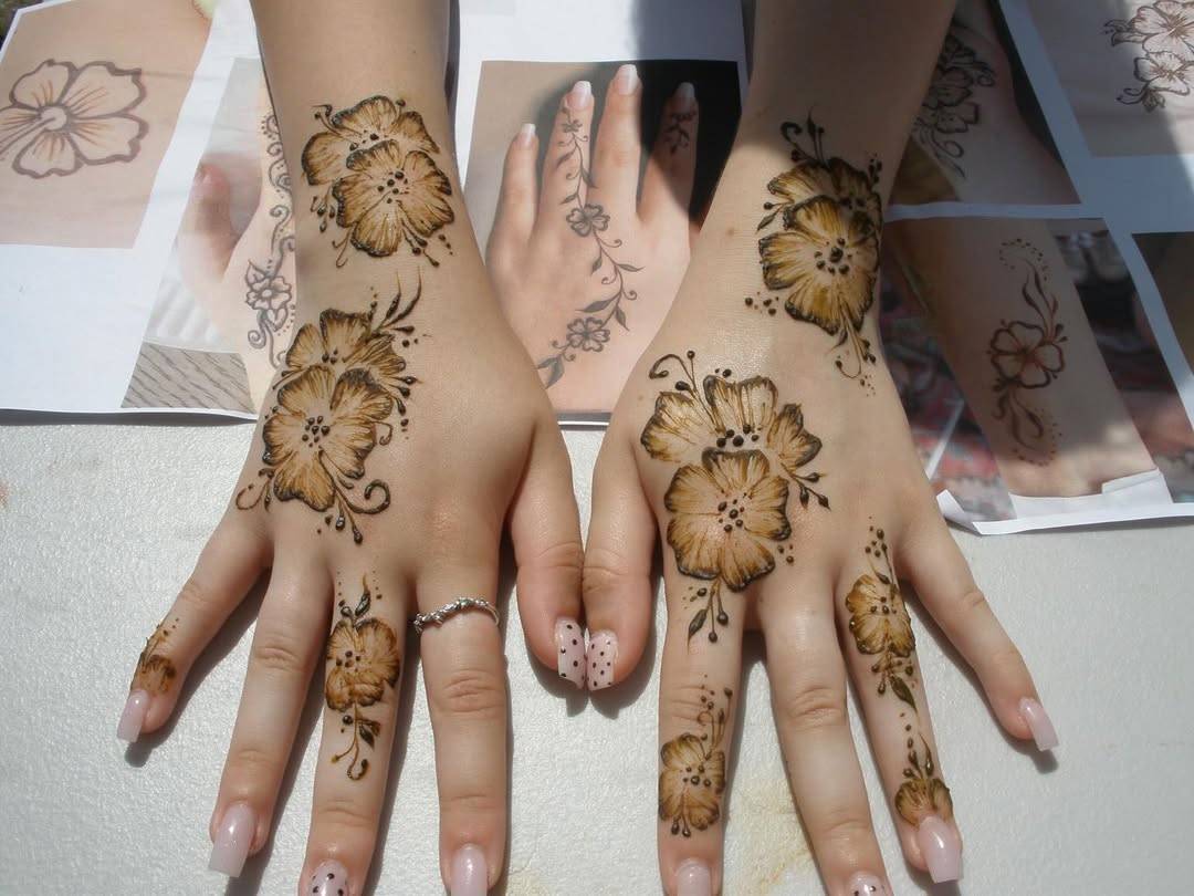 Hands adorned with brown floral mehndi extending from wrists to fingers, pale pink nails, ring on left ring finger; background shows design sheets.