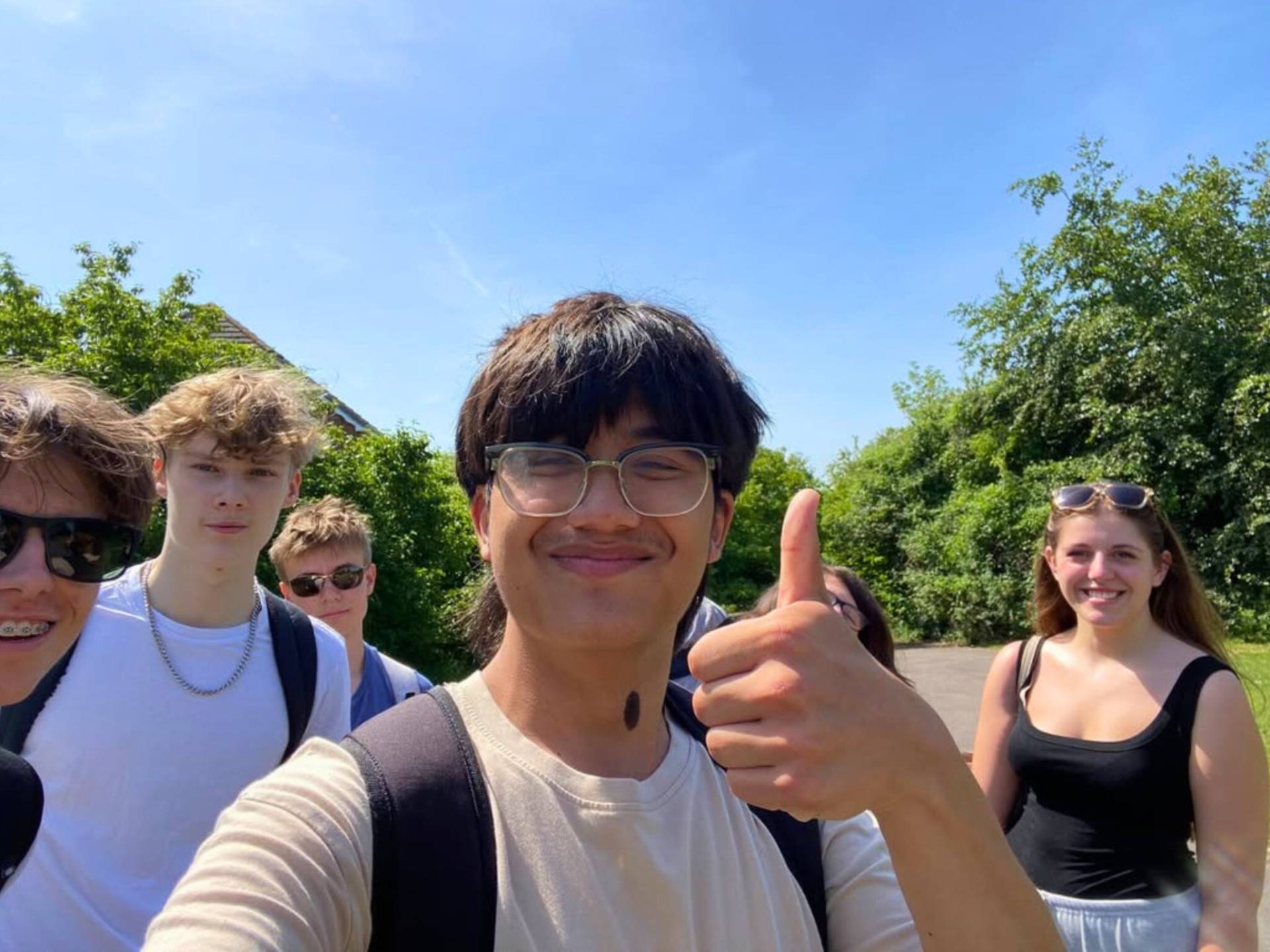 Group of friends taking a sunny outdoor selfie, smiling and standing close together with trees in the background.