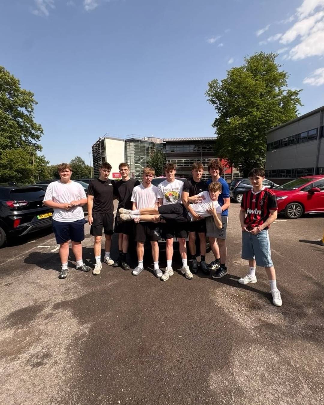 A group of young men posing in a parking lot on a sunny day, with one person carried horizontally across the arms of others in the center of the group.