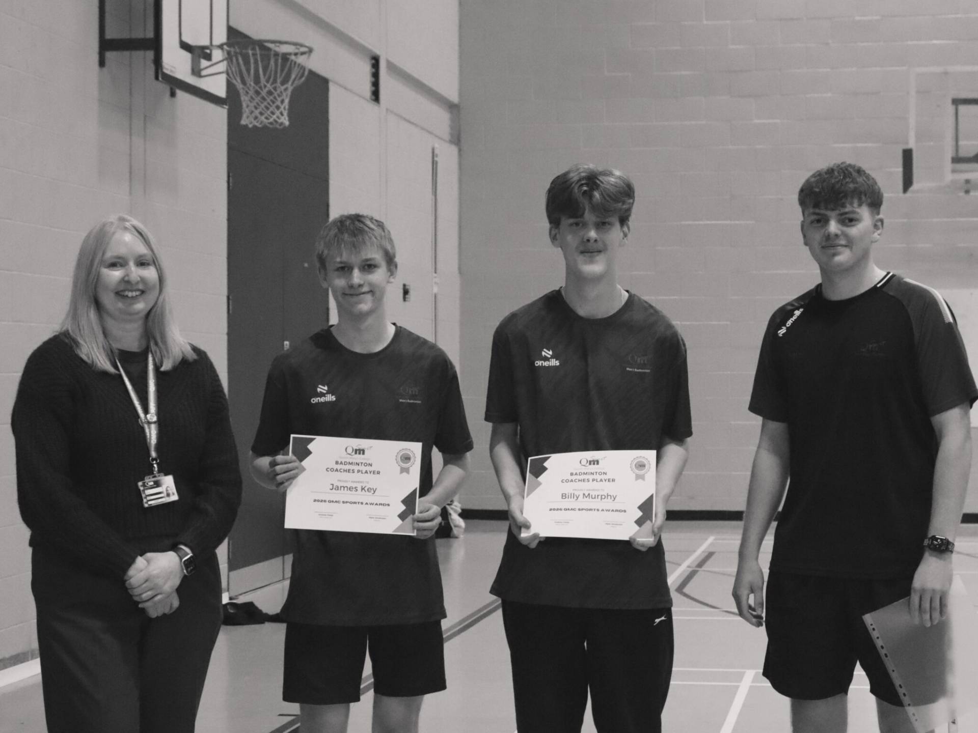 Four people pose in a gym: a woman on the left and three young men, two of whom hold certificates; basketball hoop visible in the background.