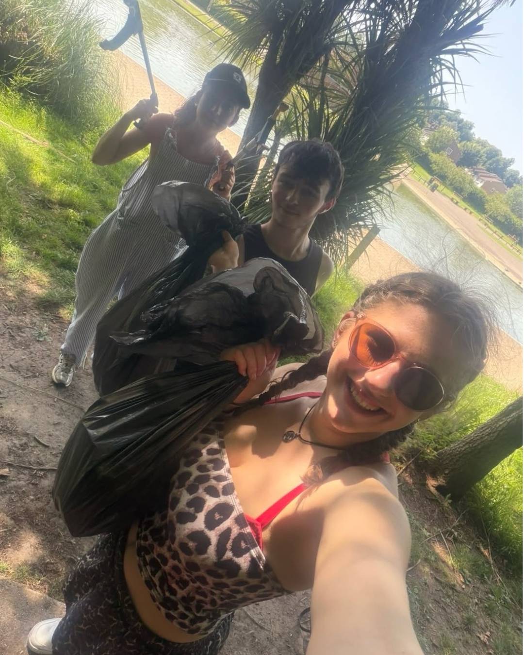 Group of friends picking up litter outdoors; one person selfies with a leopard print swimsuit top and sunglasses while others hold trash bags and grabbers near a lake.