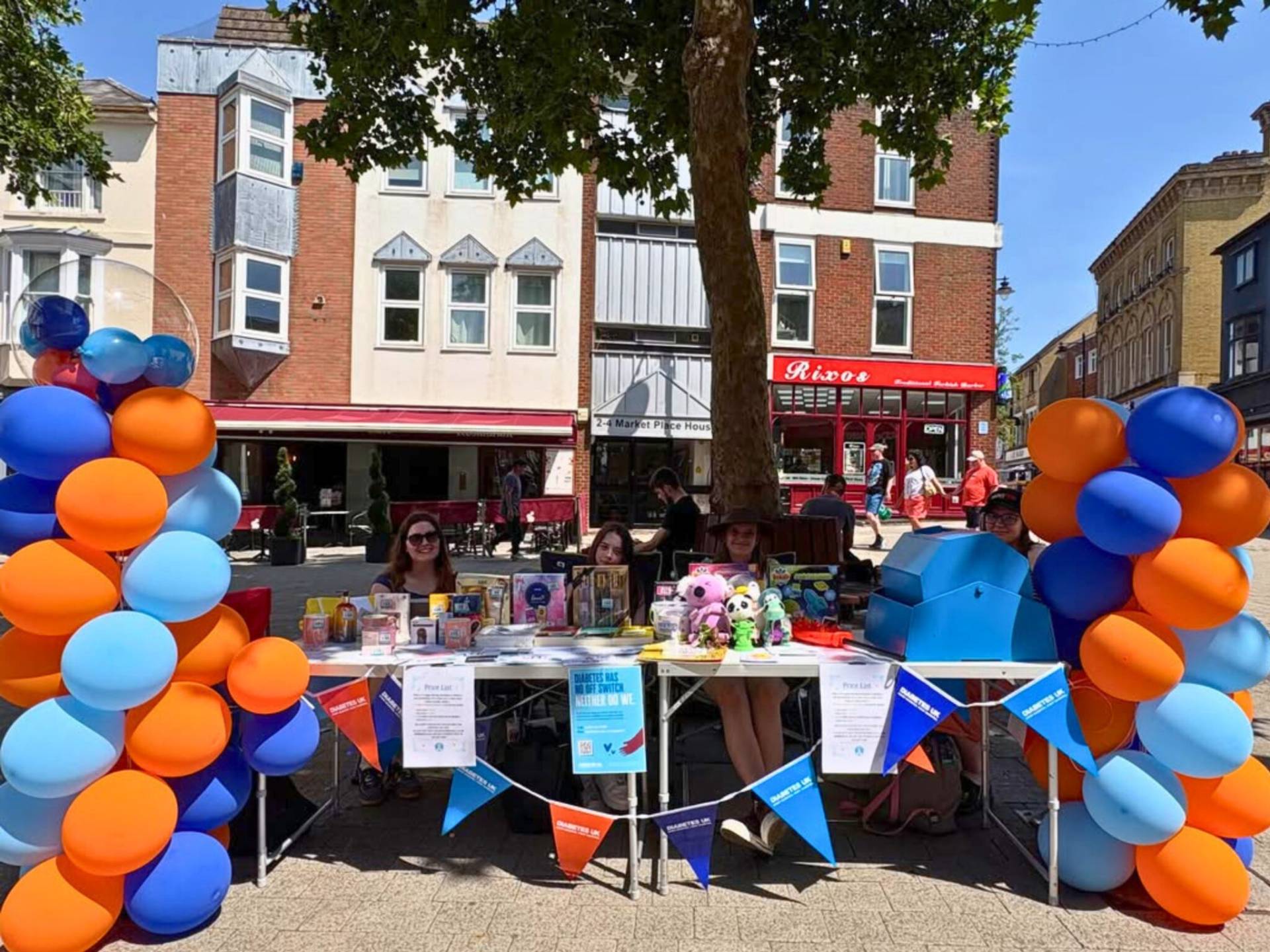 Street fair stall with orange and blue balloons surrounding a table of toys and books under a tree on a sunny day.