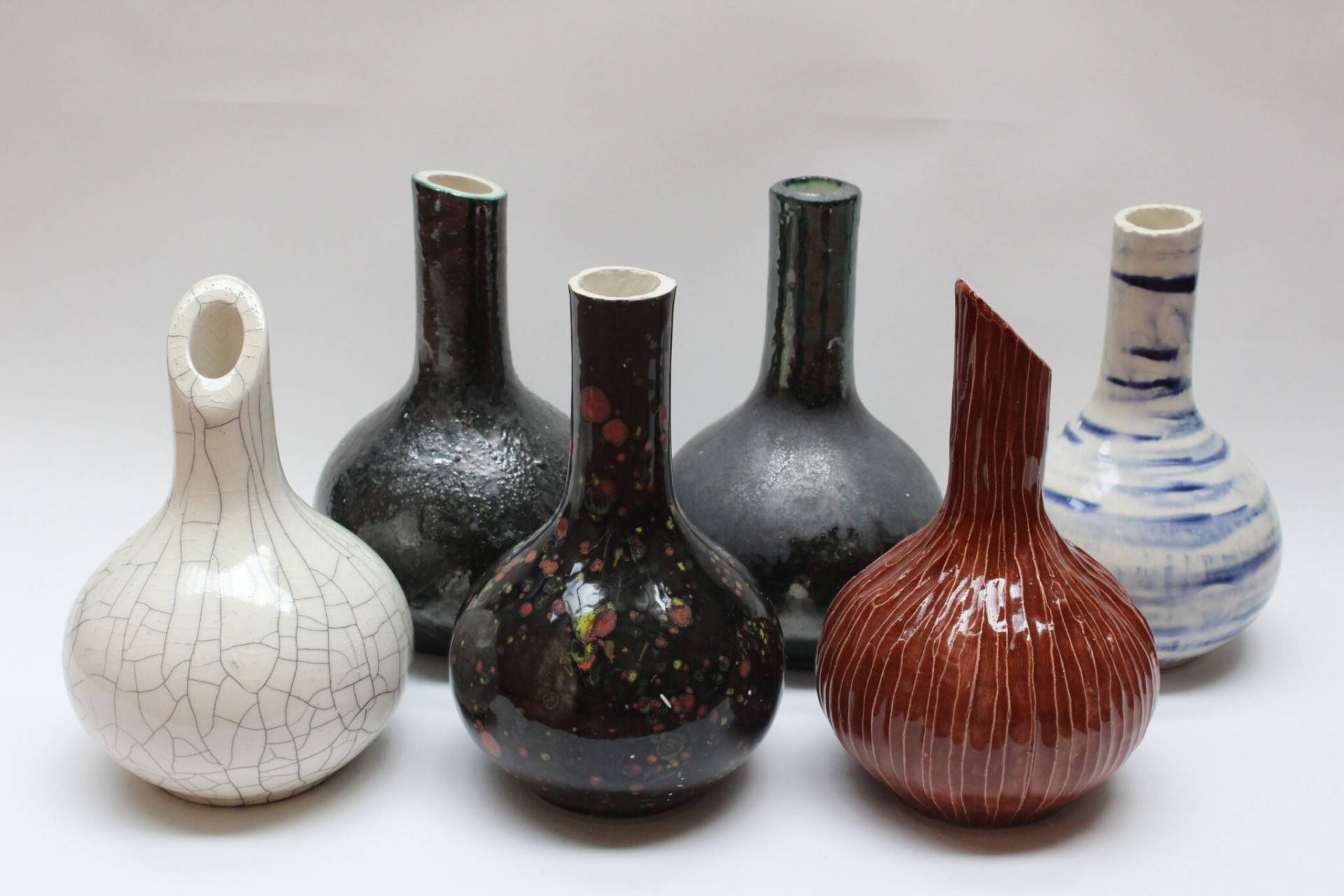 Six ceramic vases with varied glazes arranged in a row: white crackle, dark green, black speckled, gray, red ribbed, and blue-striped.