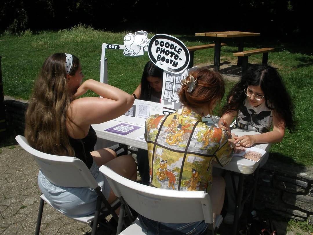 Four people sit around a white DIY photo booth table outdoors, with a circular 'Doodle Photo Booth' sign visible nearby.