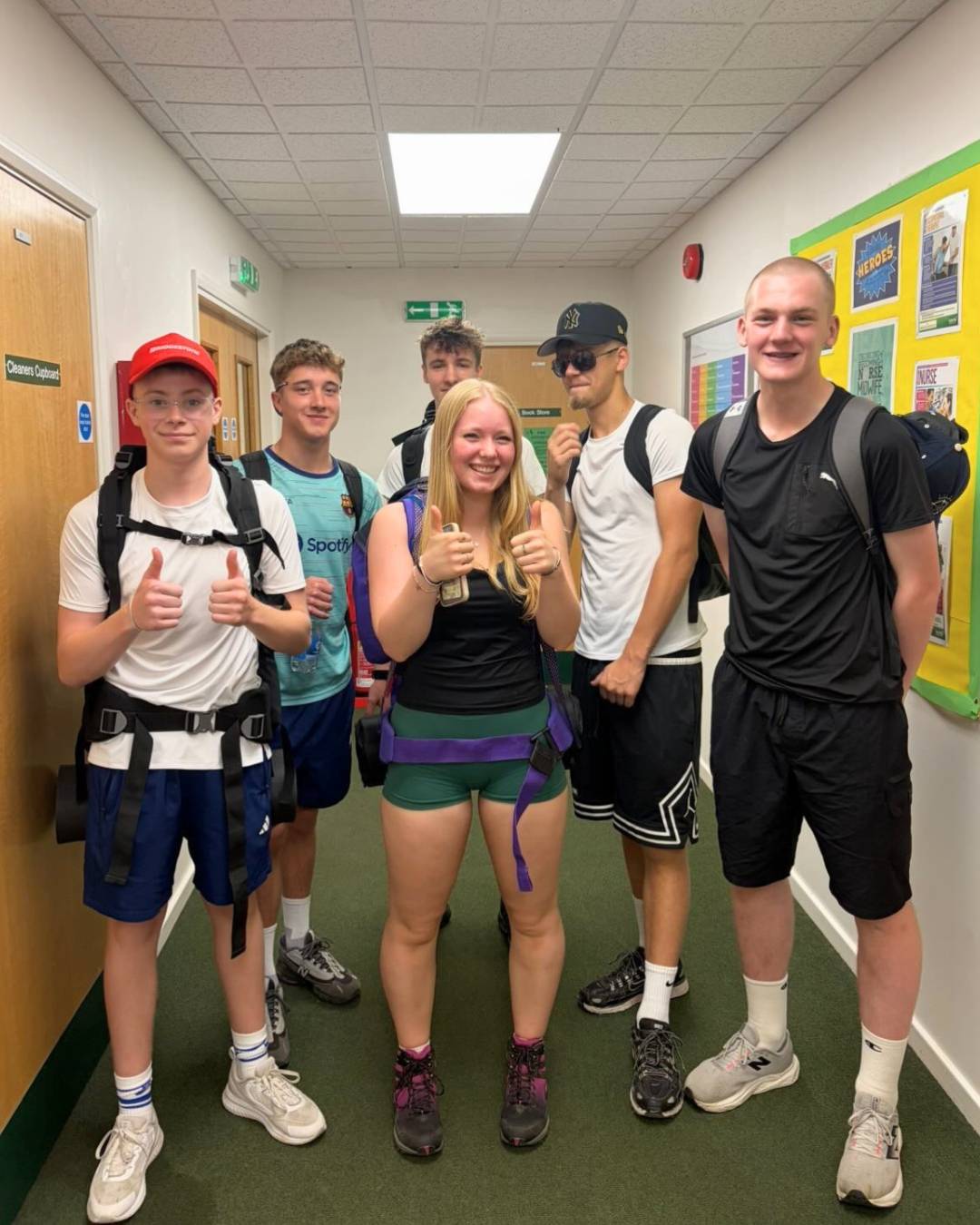Six teenagers in a school hallway with backpacks, posing and giving thumbs up to the camera.