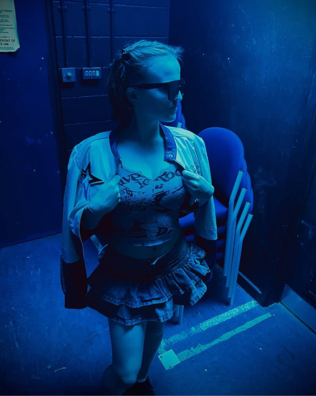 Person wearing sunglasses, an open jacket over a graphic crop top and ruffled skirt, posed in a blue-lit backstage area.