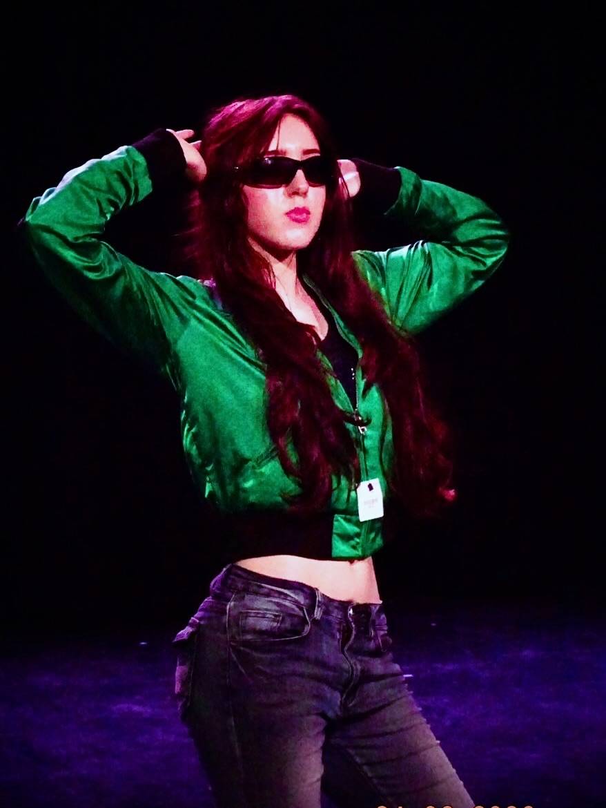 Woman performer in a green cropped jacket and sunglasses raises her arms on a dark stage.