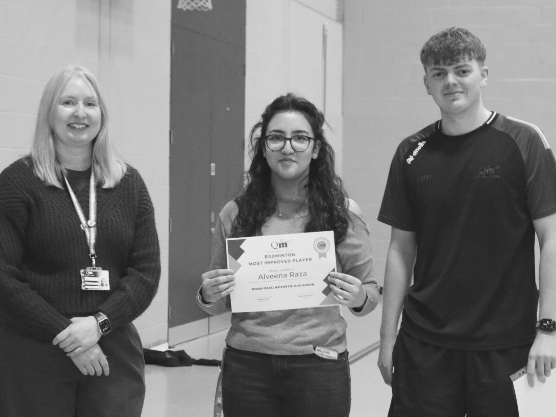Three people posing together in a hallway; center person holds a certificate for sports award, flanked by a smiling woman on the left and a man on the right.