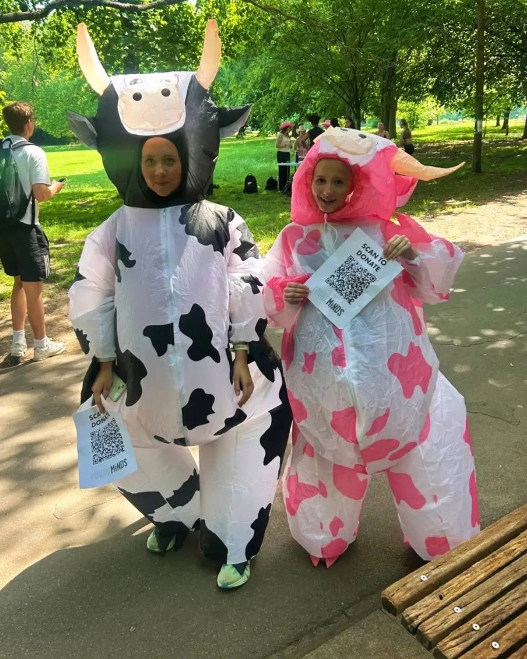 Two people in inflatable cow costumes (black-and-white and pink-and-white) pose in a park, each holding a QR code flyer.