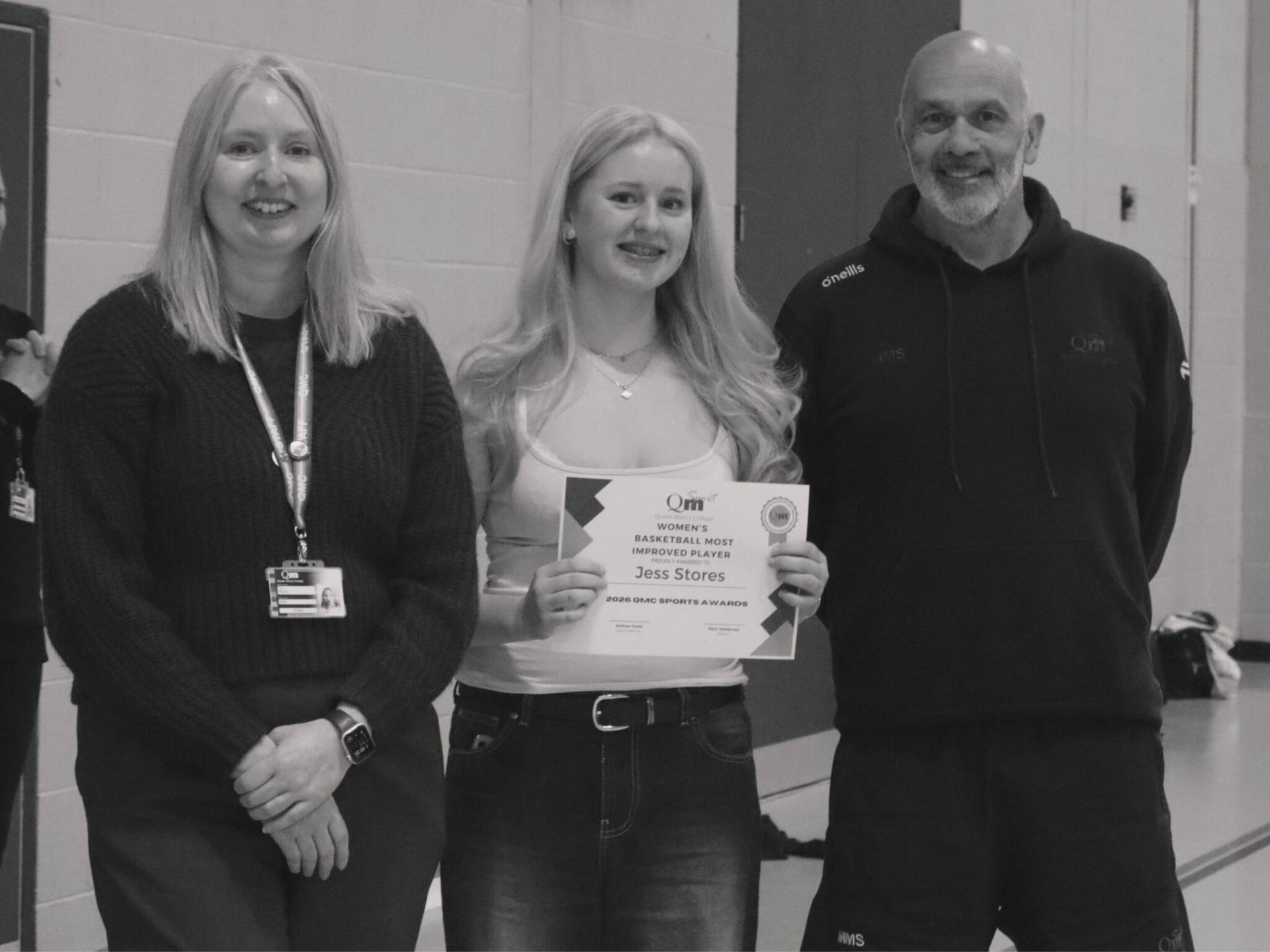 Three people posing indoors; woman in middle holds a certificate and smiles, flanked by a woman left with lanyard and a man on the right in a hoodie.