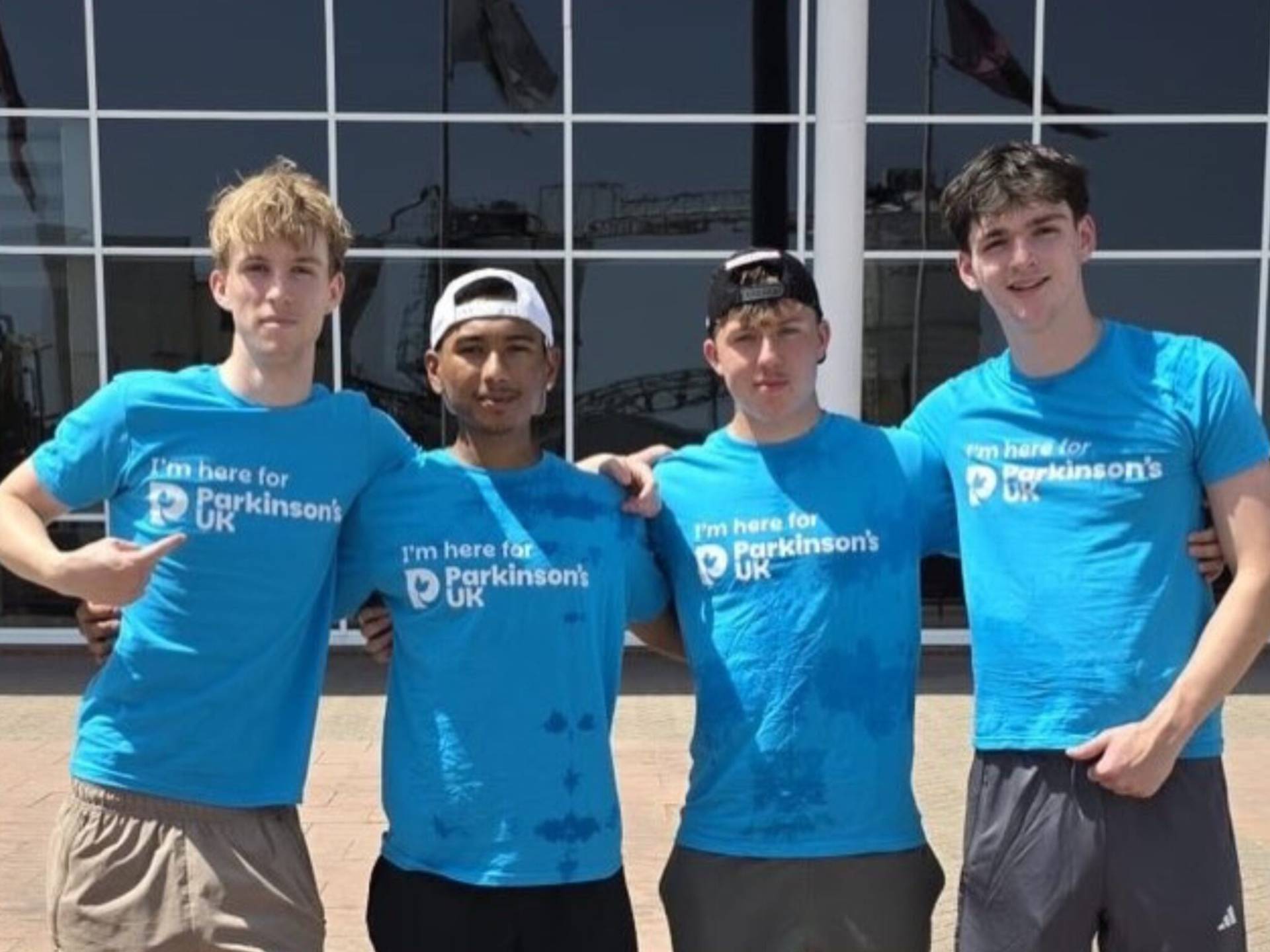 Four young men in blue Parkinson's UK shirts stand with arms around each other outside a glass-front building.