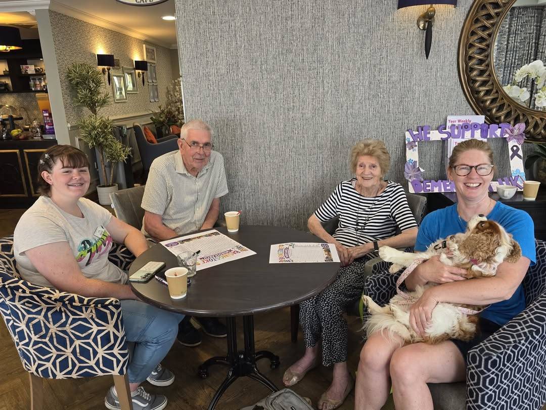 Four people sit around a round cafe table, smiling; a woman on the right holds a fluffy dog.