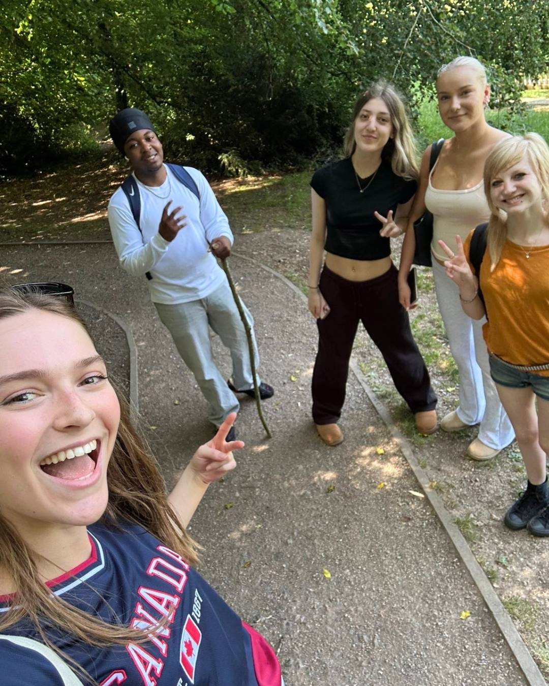 Group of five young adults posing for a selfie on a gravel park path, with trees in the background; everyone smiles and shows peace signs. The person in the foreground wears a Canada jersey.