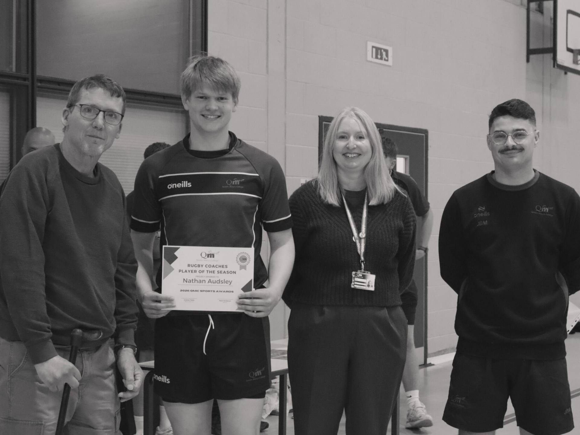 Group of four people in a gym: a young man in sportswear holds a certificate, flanked by smiling adults, at an awards event.