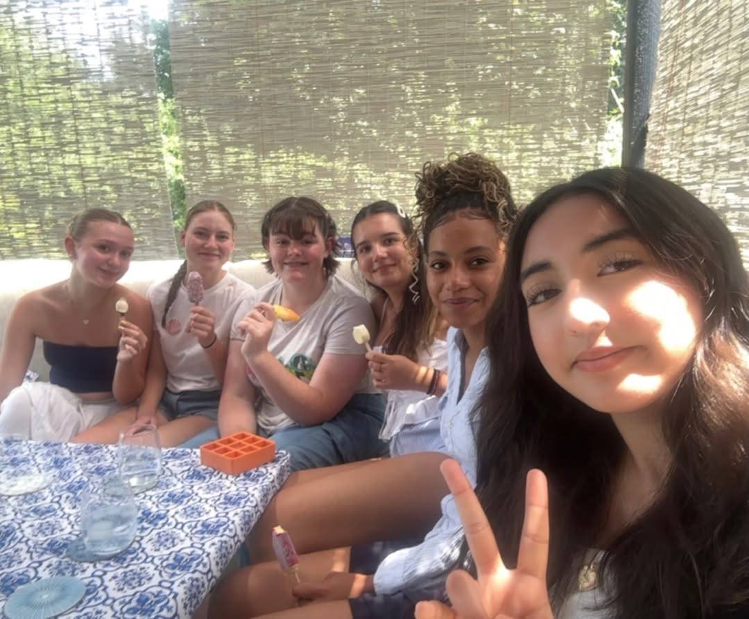 Six friends sit together under a shaded patio, each holding colorful ice pops and smiling at the camera during a sunny outdoor gathering.