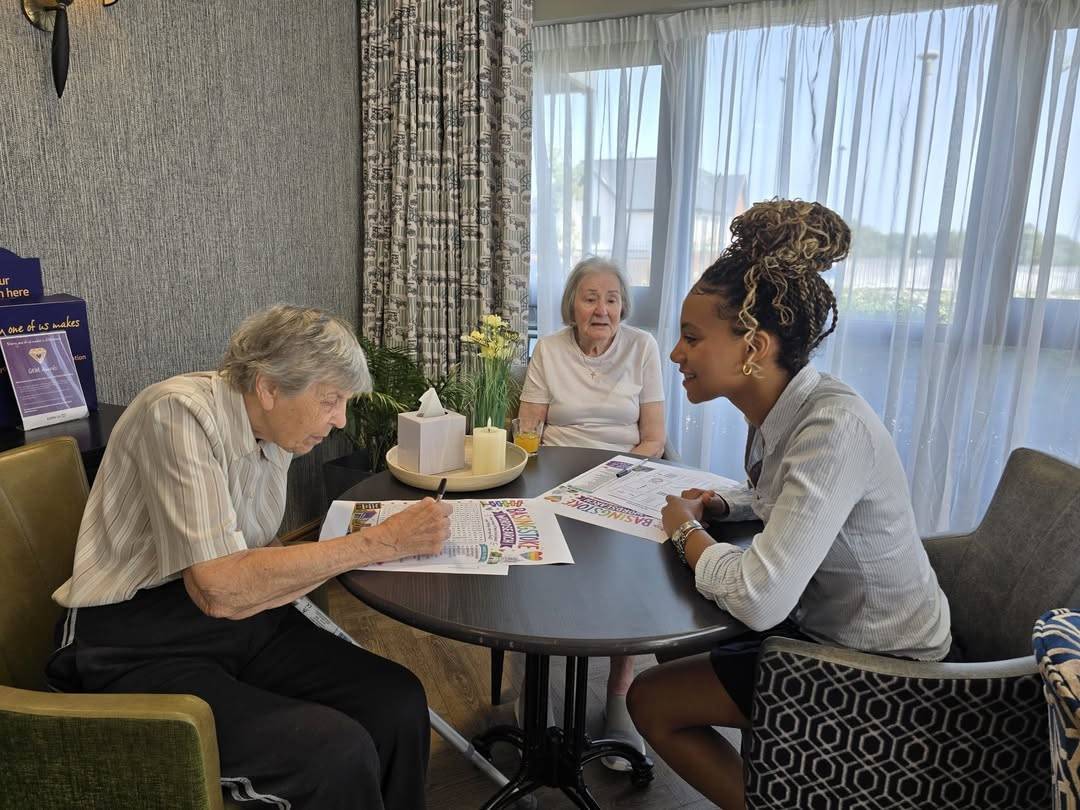 Three women sit around a round table in a bright room; an elderly woman on the left writes on a colorful flyer while two other women converse, with a tissue box, candles, and flowers on a tray in the center.