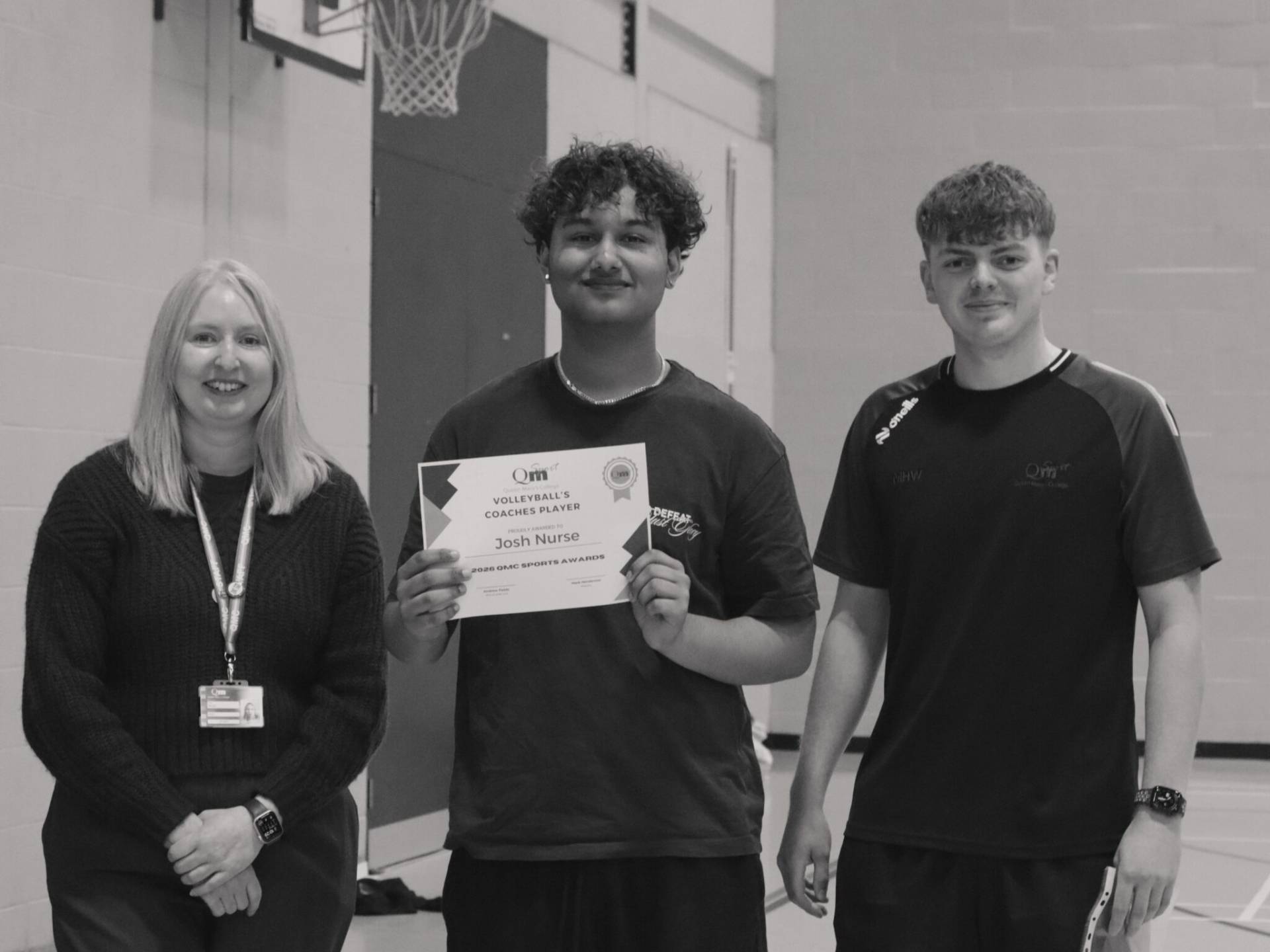 Three people stand in a gym posing for a photo; the central person holds a certificate while the other two smile beside them.
