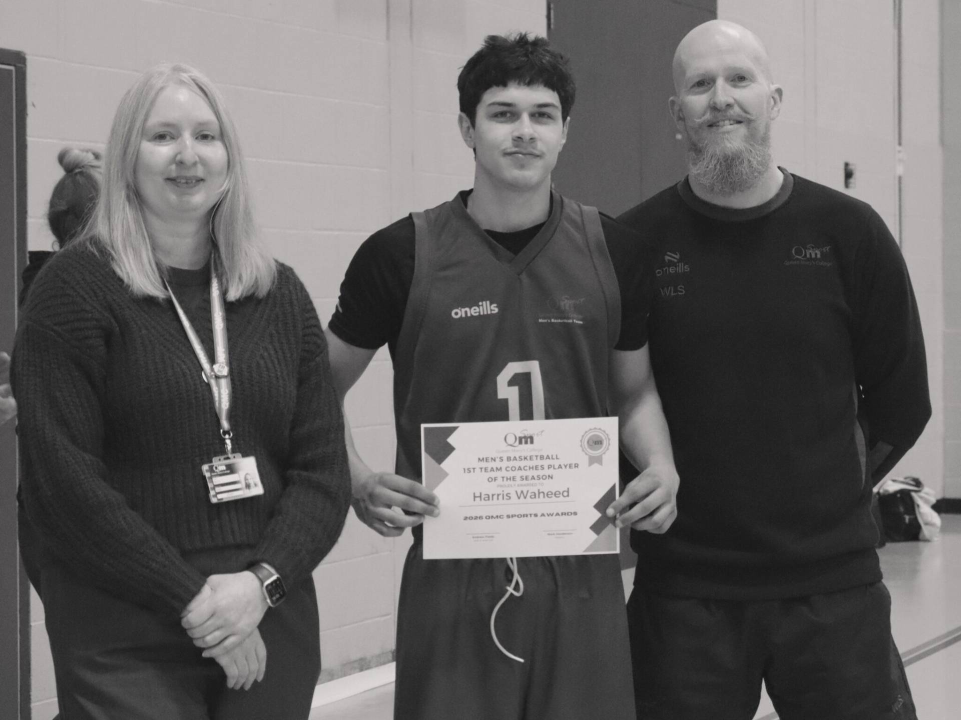 Three people posing for a group photo at an awards event; the center athlete in a basketball jersey holds a certificate while flanked by a woman and a man in a gymnasium setting.