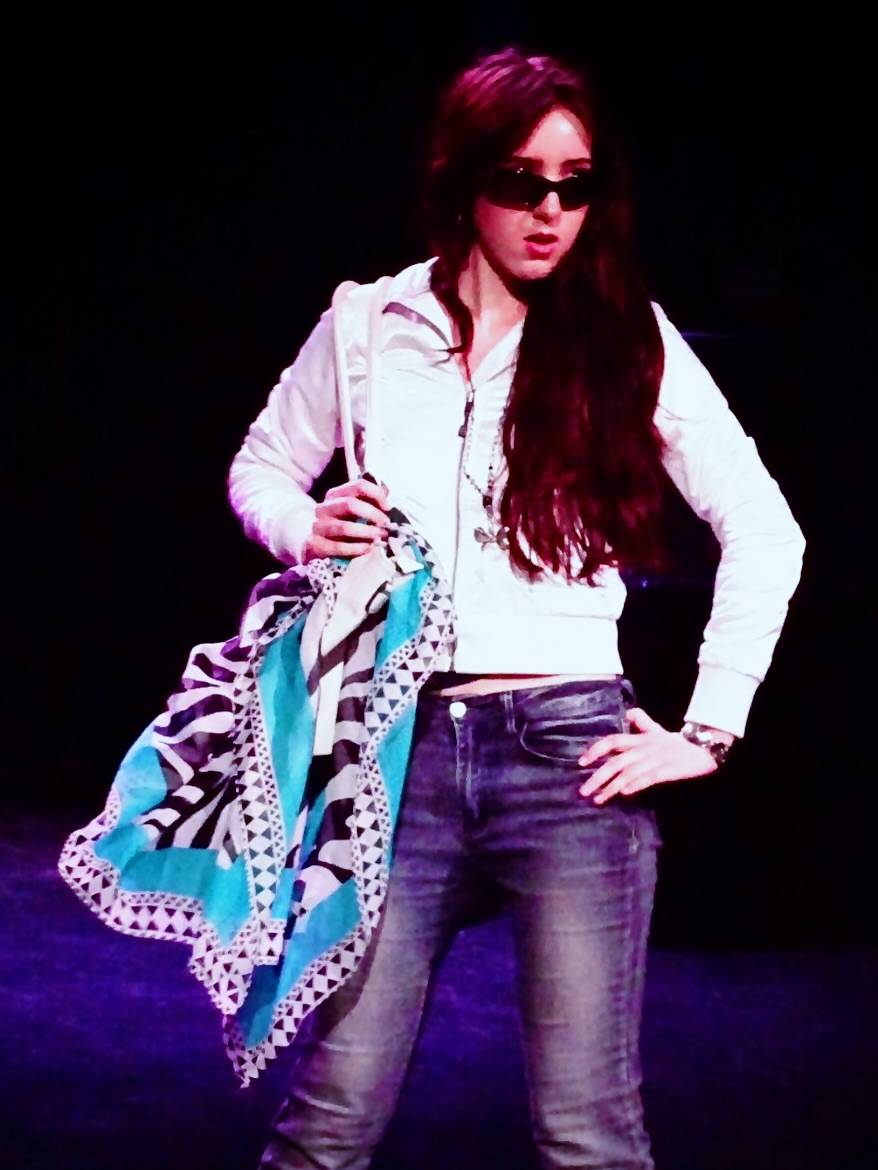 Woman on stage in a white jacket and jeans, wearing sunglasses and holding a blue patterned scarf on her shoulder.