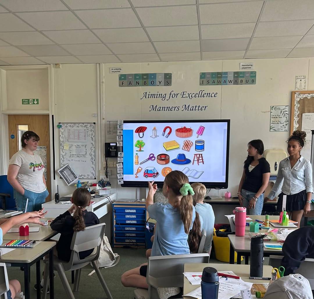 Classroom scene: students watch a large screen showing food-item icons during a lesson, with teachers at the front and a banner reading 'Aiming for Excellence, Manners Matter' on the wall.