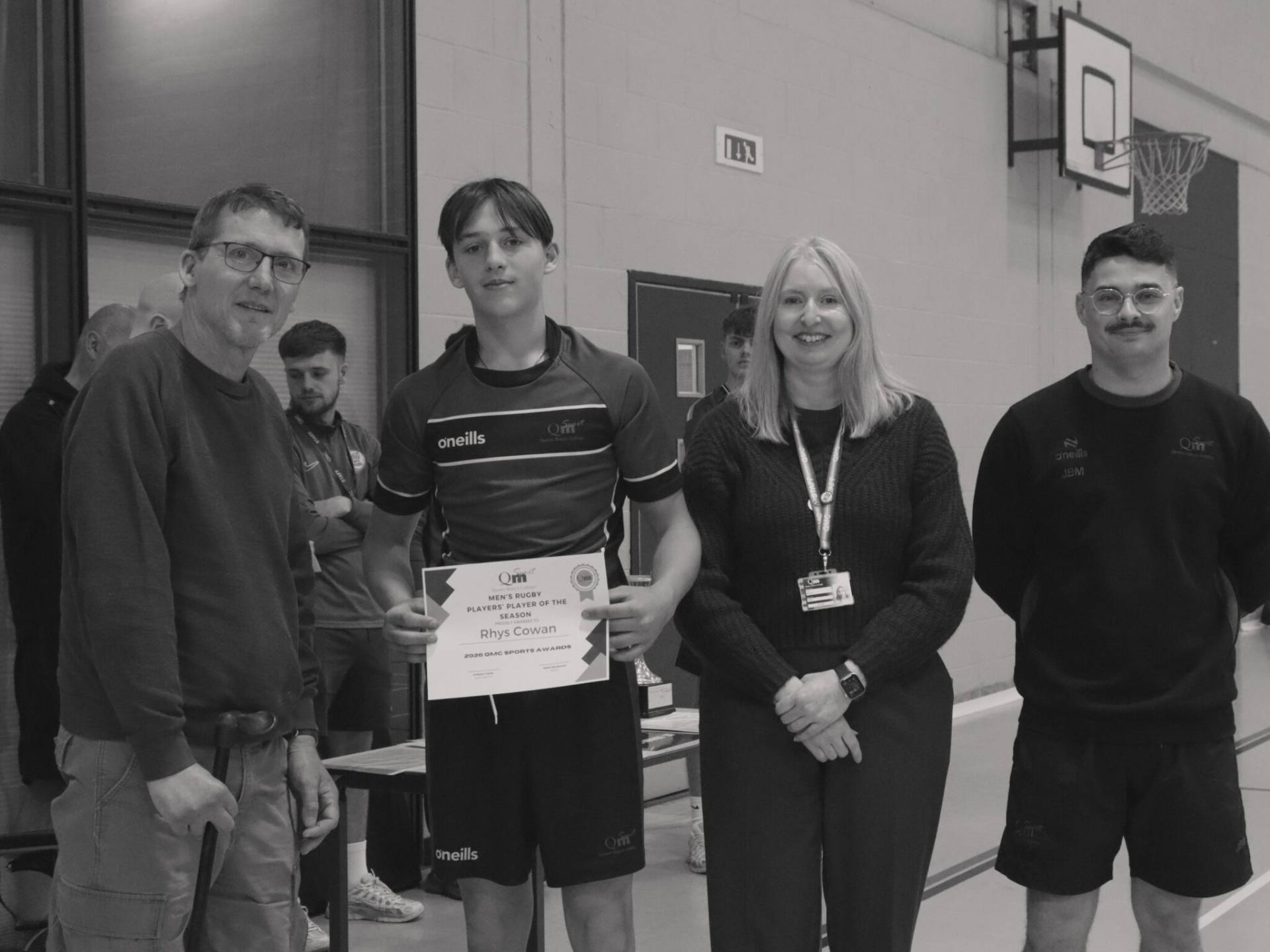 Group of people in a gym posing for a photo; a young man in a sports shirt holds a certificate, flanked by teammates and others smiling in the background.