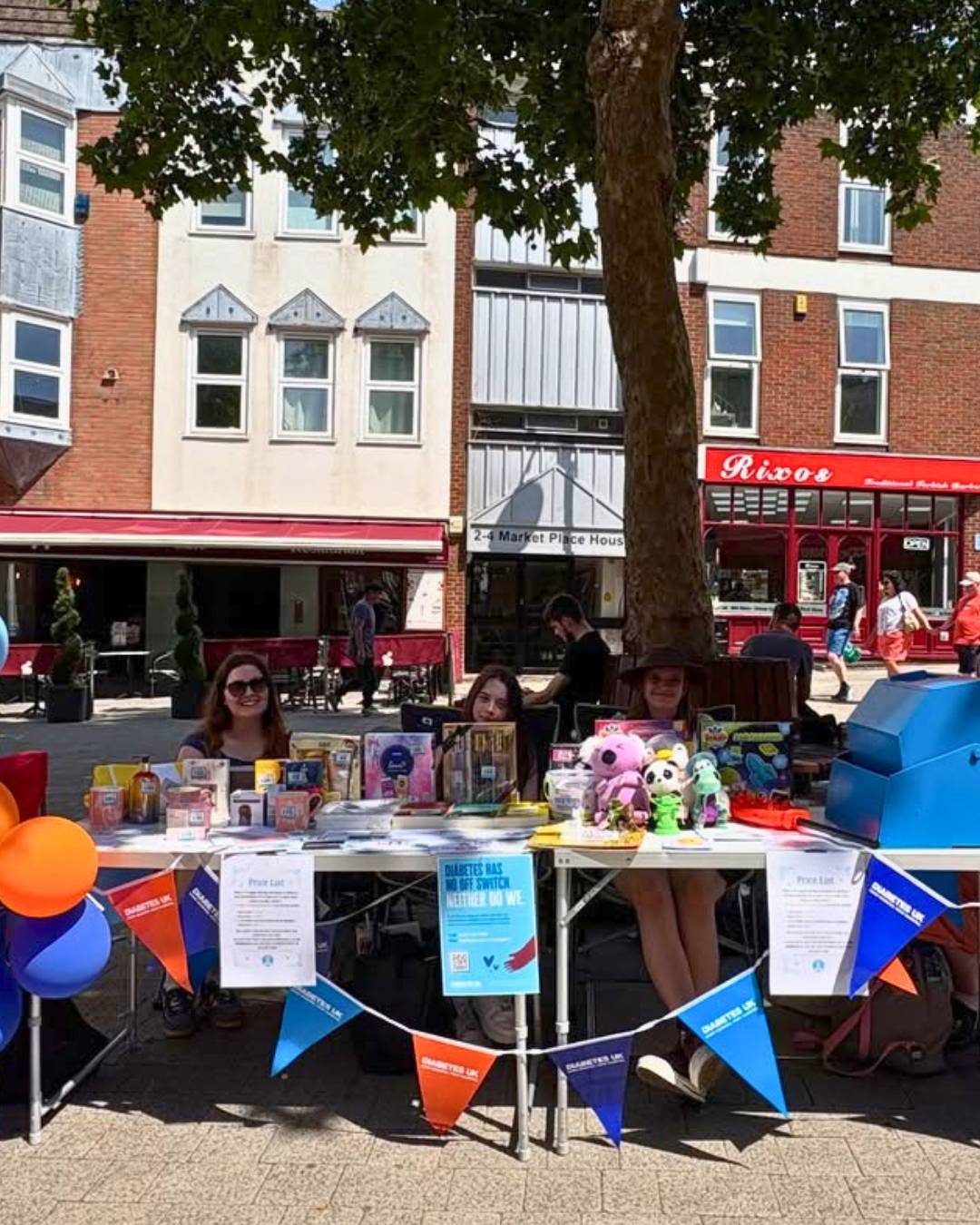People sit at a street table selling books and toys with blue/orange bunting; a small crowd and shops in the background.