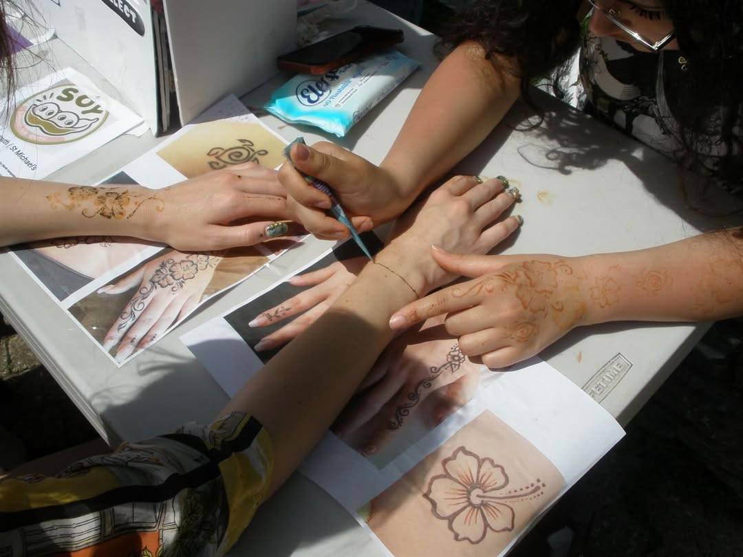 Group of people applying henna on each other's hands at a table with stencil sheets nearby at a workshop.