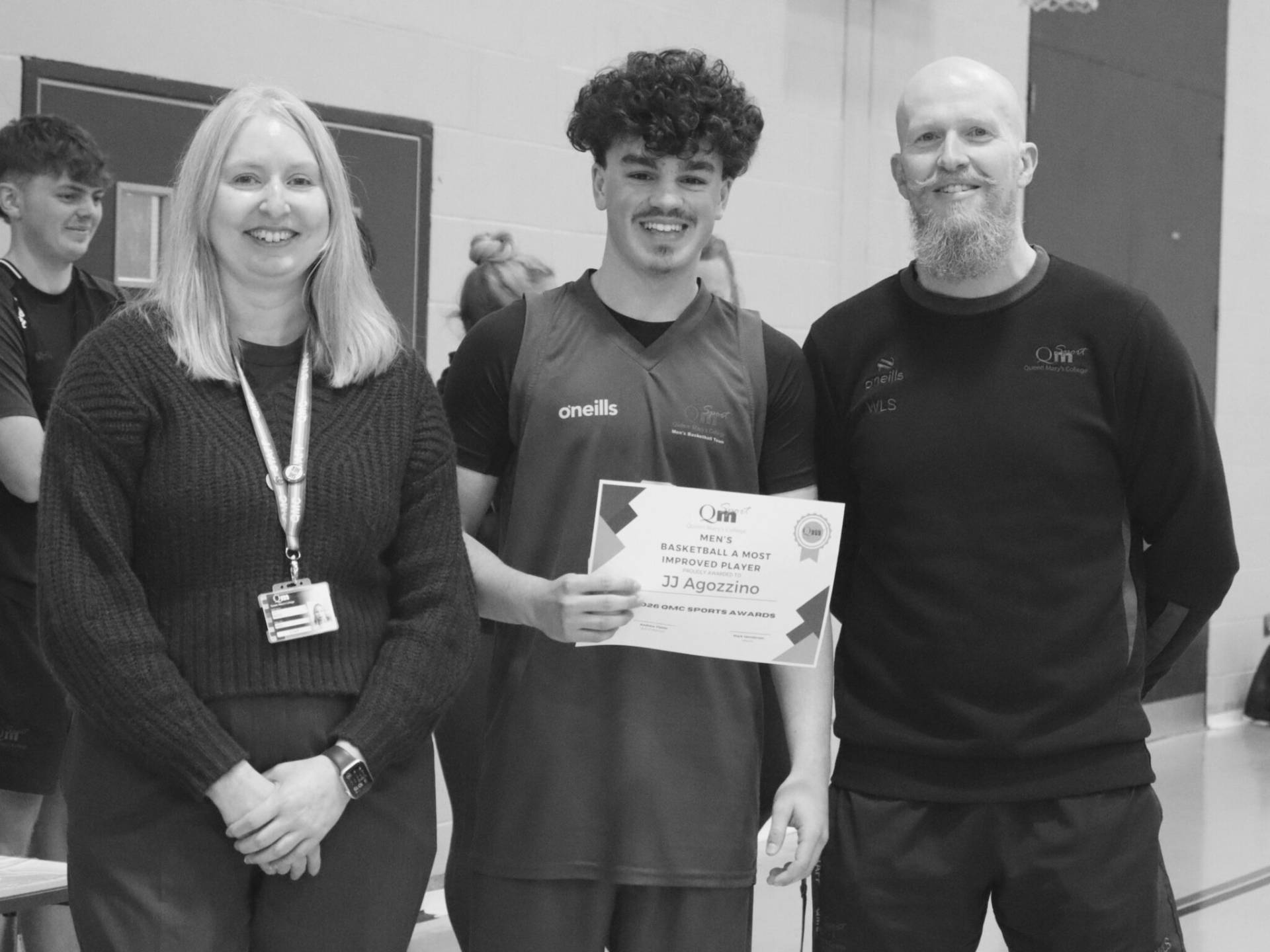 Three smiling people indoors: woman with lanyard left, man in jersey center holding a certificate, bearded man right.