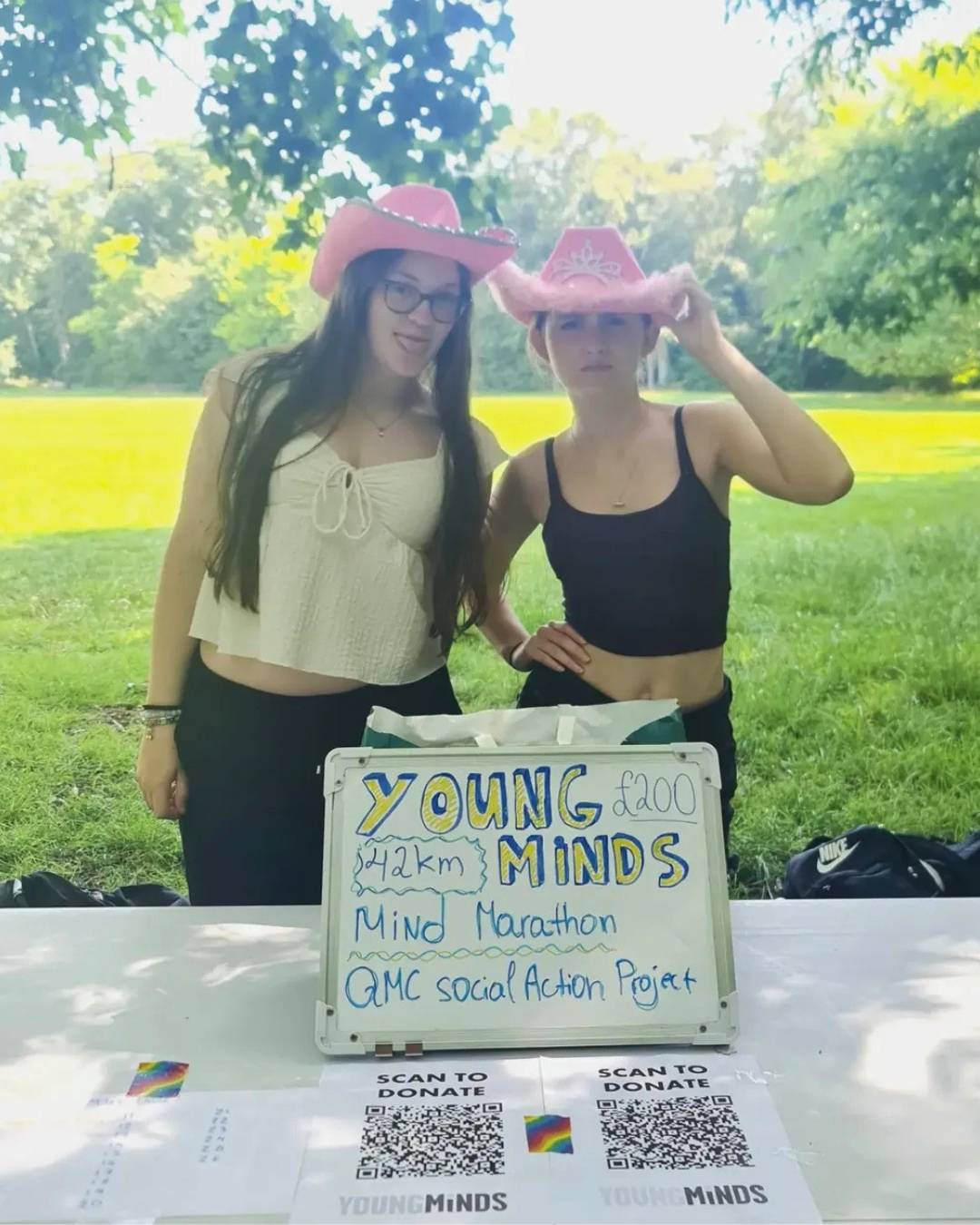 Two young women in a park wearing pink cowboy hats pose beside a sign for the Young Minds Mind Marathon fundraiser. Common, cheerful outdoor charity scene.