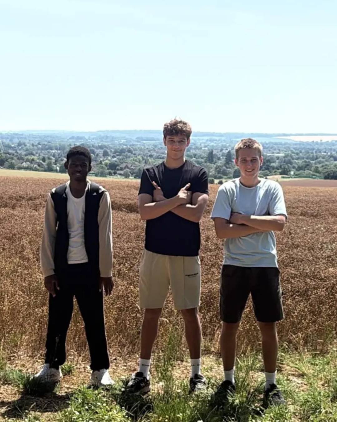 Three young men stand in a brown field on a sunny day, arms crossed, overlooking a rural landscape.
