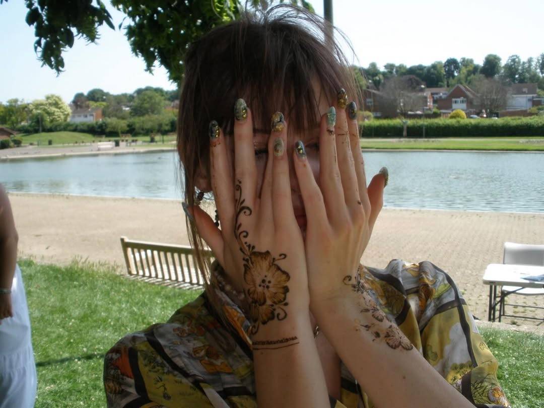Person outdoors with hands raised to partially cover their face, displaying intricate henna designs on both hands and forearms.
