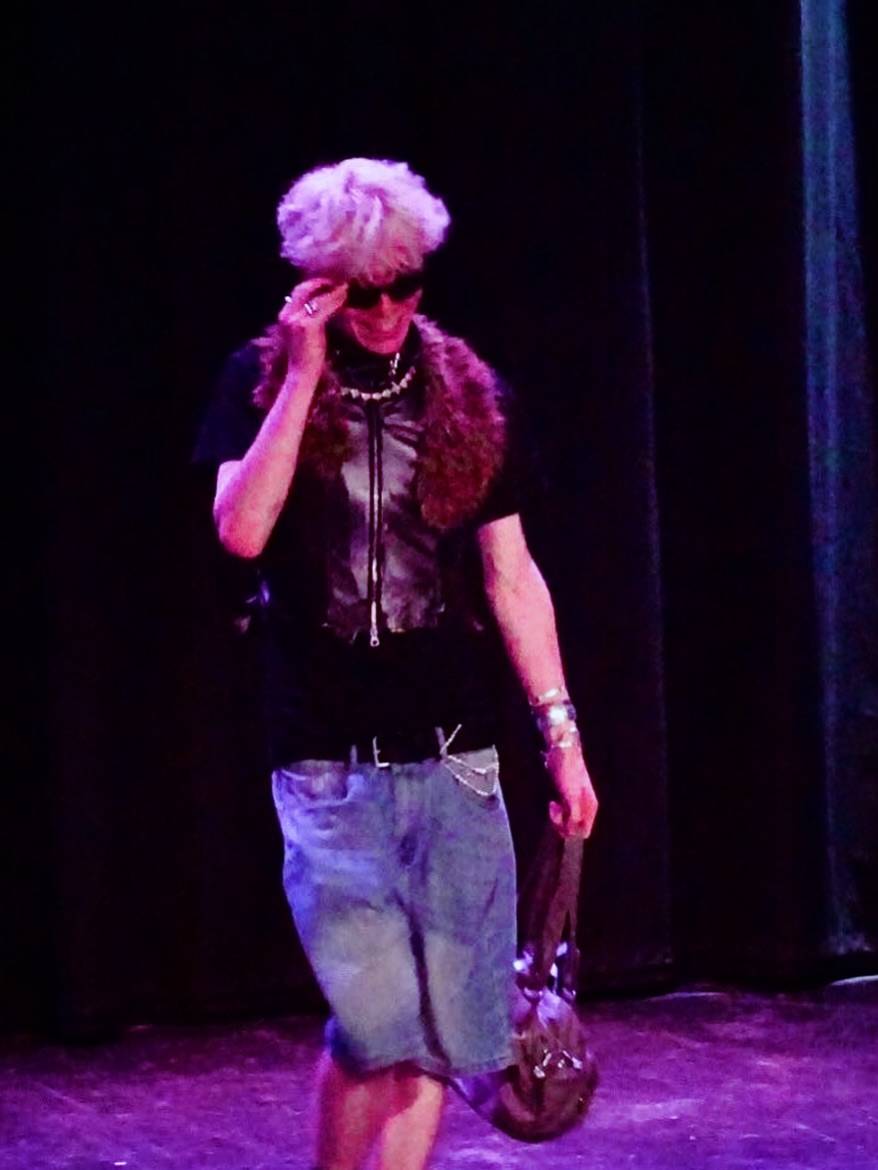 Person on a dimly lit stage wearing sunglasses, a dark vest, denim shorts, and a furry scarf, holding a brown bag or purse.