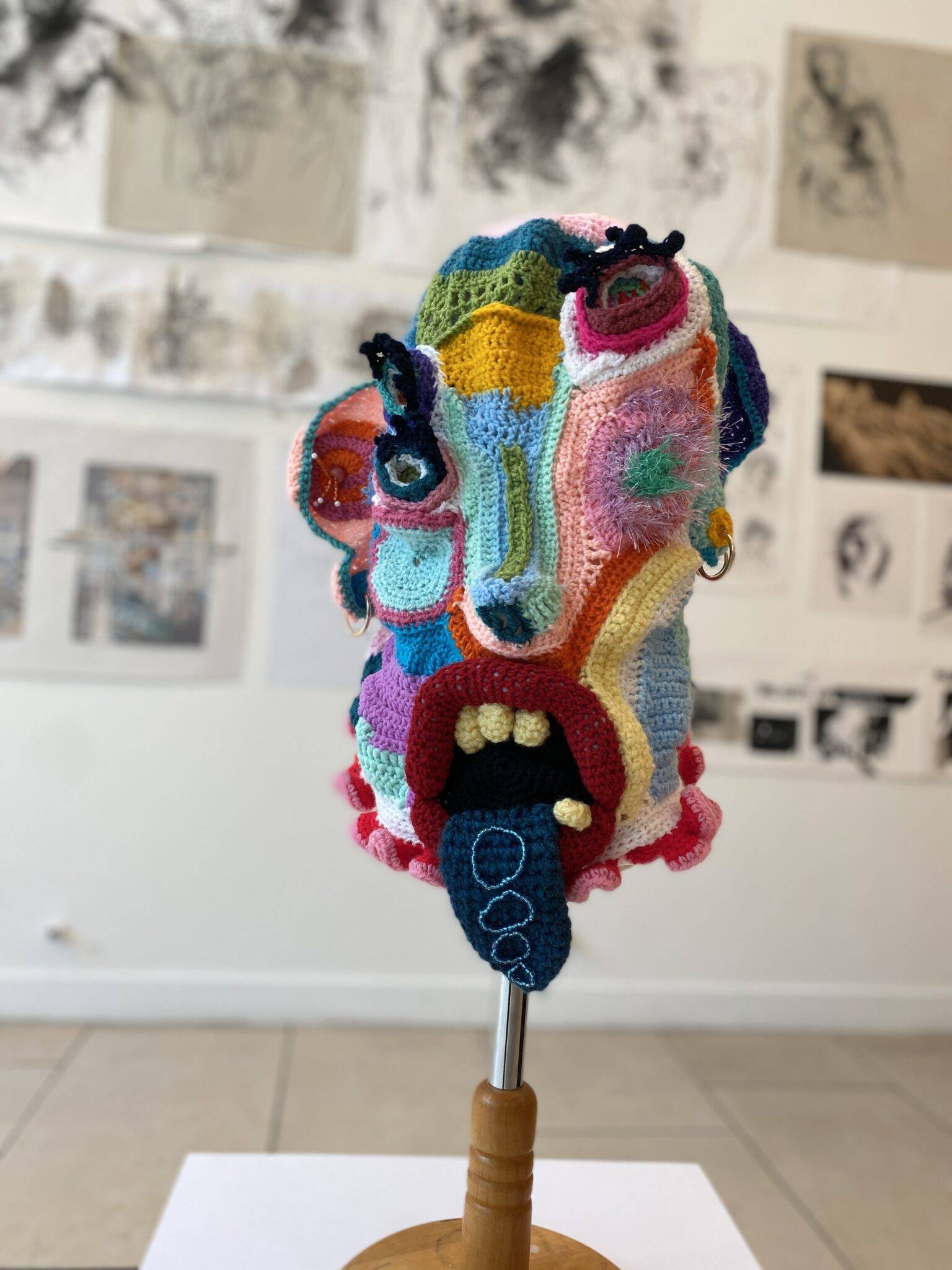 Colorful crocheted sculpture of a distorted head made from multicolored patches, displayed on a wooden stand in a gallery setting.