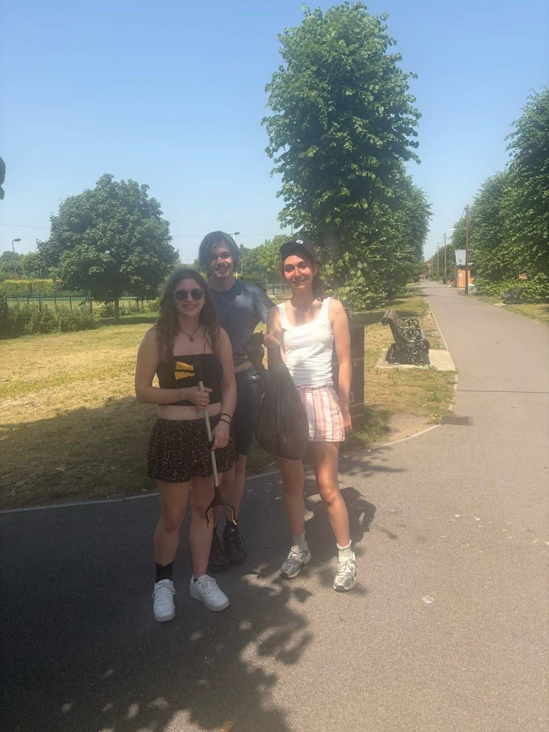 Three friends posing outdoors on a sunny day; one person holds a litter picker and another carries a black trash bag.
