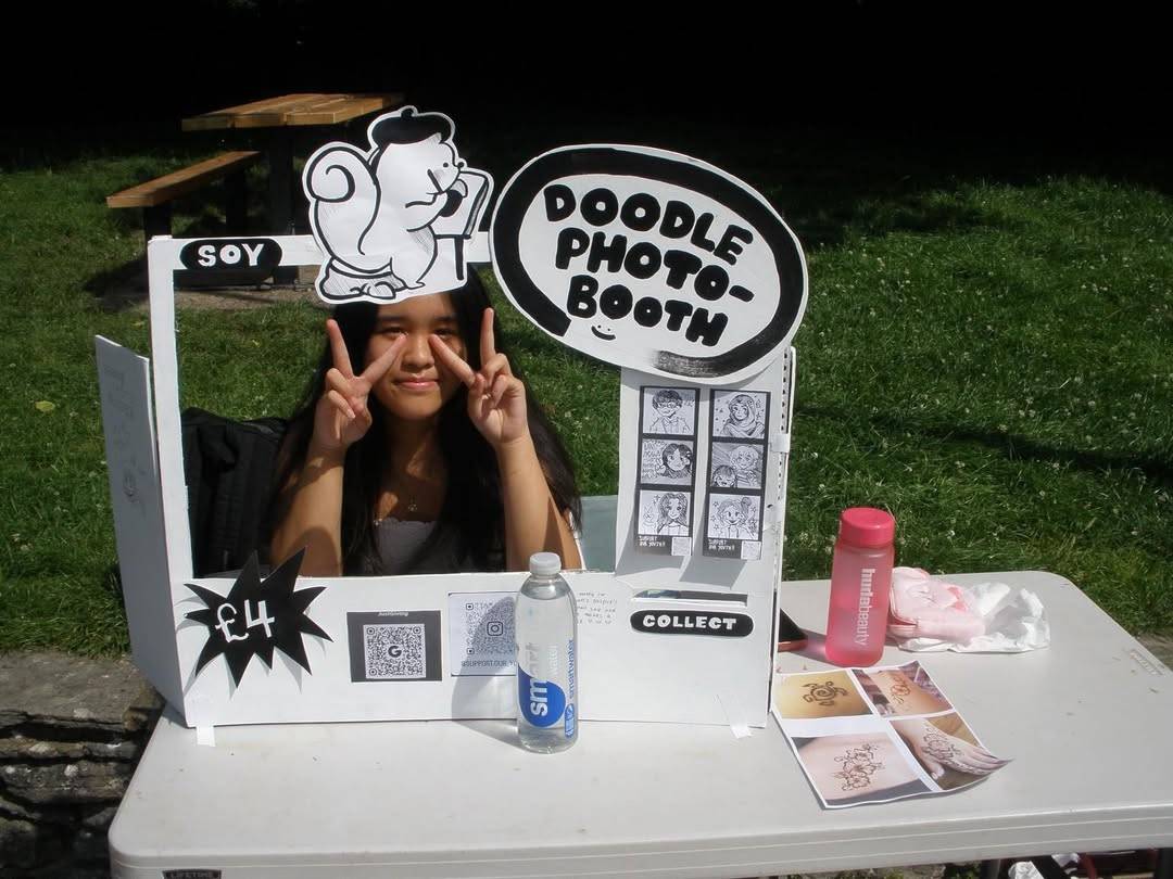 Girl in a white DIY photo booth at a park, making peace signs with both hands as the sign reads 'Doodle Photo Booth' and '£4' on the side.
