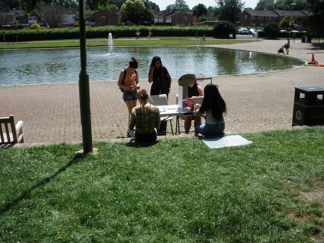 Group of five people gathered around a small folding table on a grassy hillside by a circular pond with a fountain, doing crafts in a sunny park.