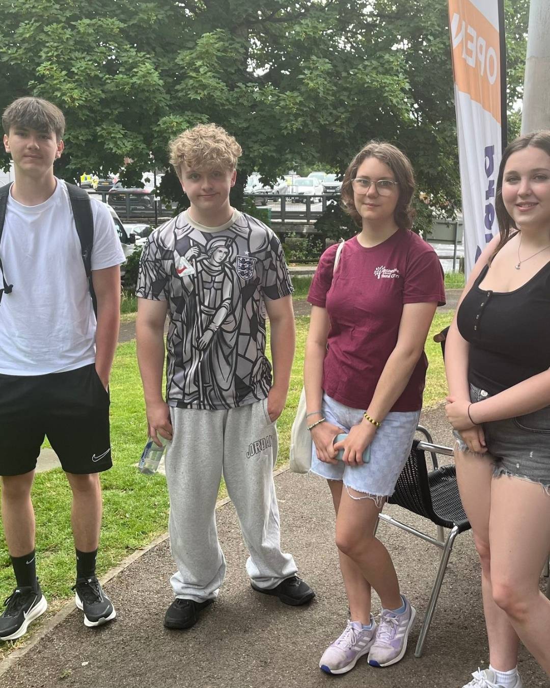 Group of four teenagers standing on a sidewalk outdoors, posing for a photo with green grass and trees in the background and a tall orange-and-white banner to the right.