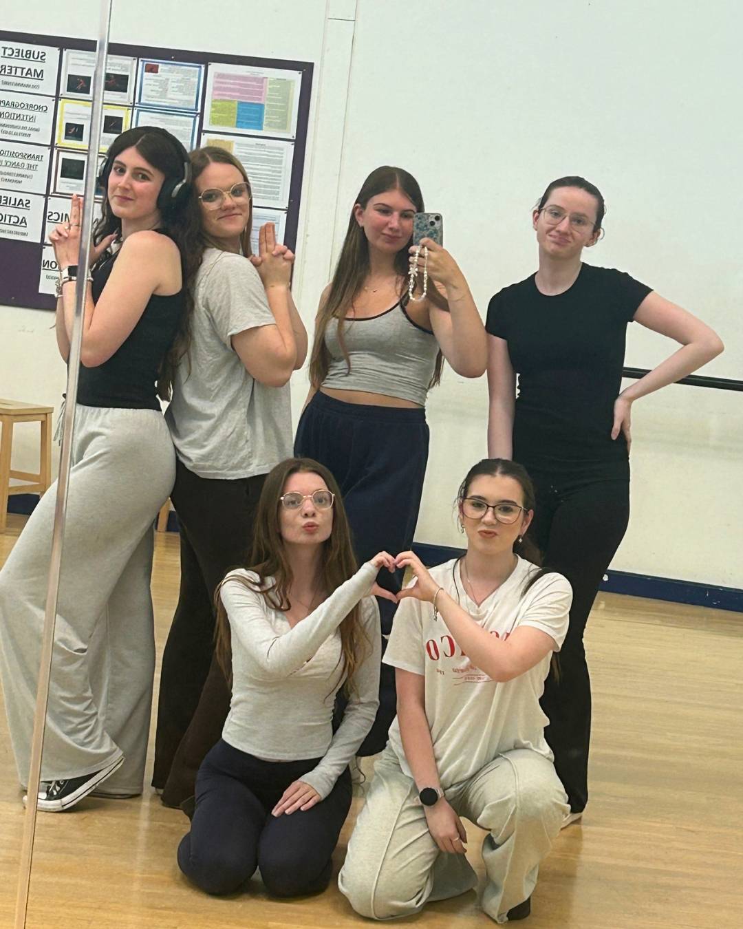Six young women in a dance studio pose for a mirror selfie; two kneel in front making a heart with their hands.