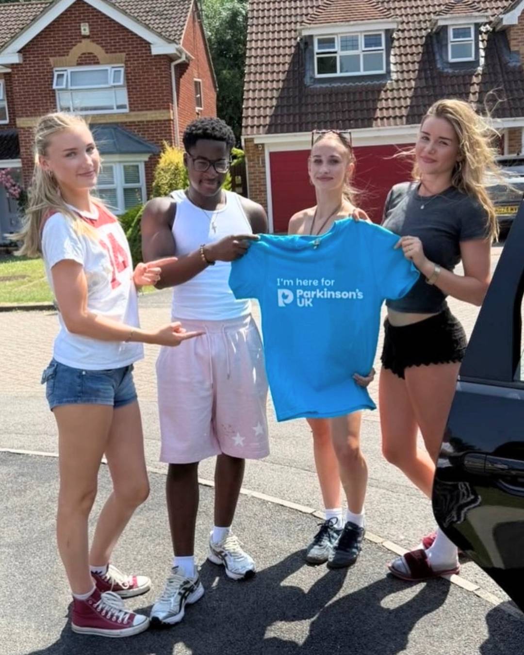 Four young adults pose on a residential street, holding up a blue Parkinson's UK t-shirt.
