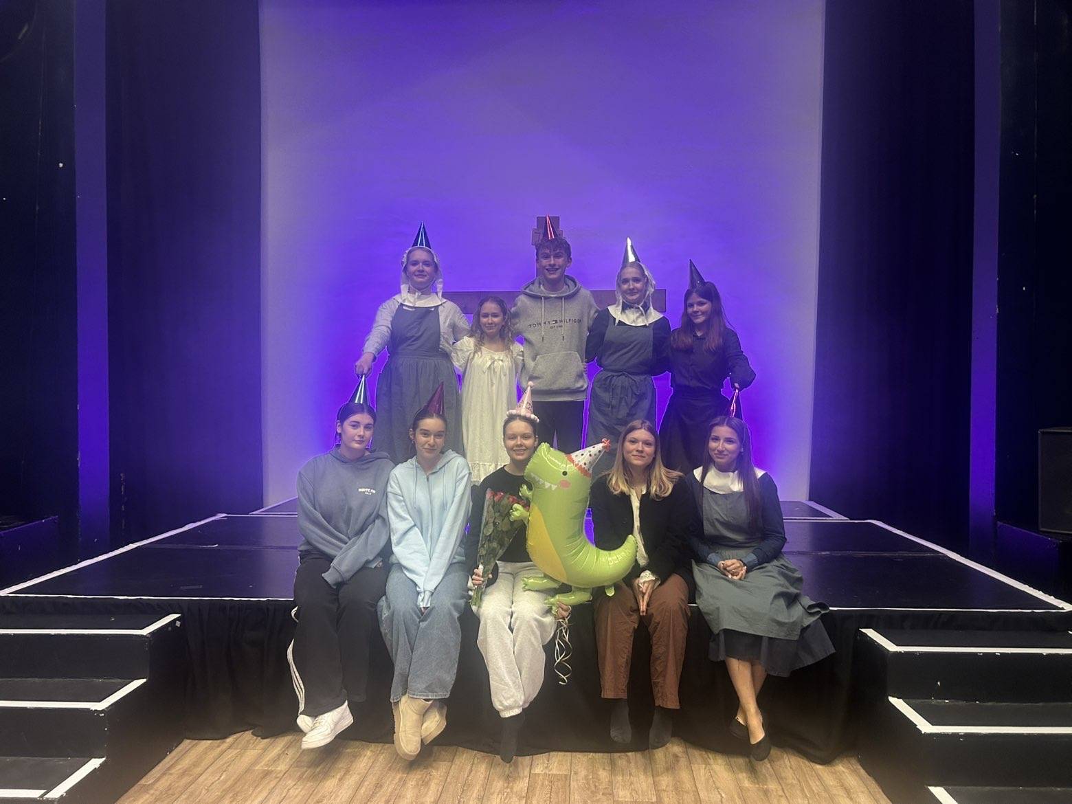 Ten young people on a theater stage wearing party hats, posing for a group photo; one holds a green dragon balloon.